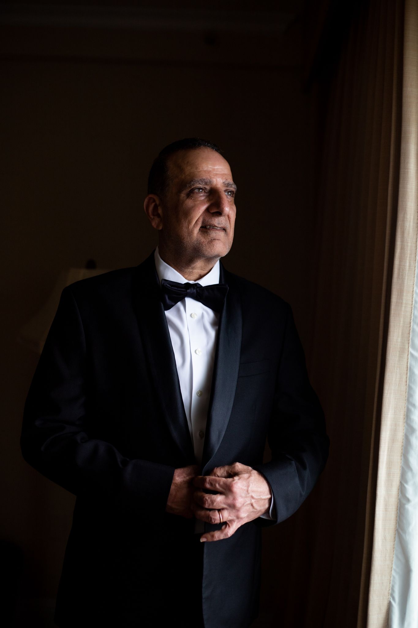 Elegant older man in black tuxedo and bow tie standing by a window in dramatic lighting.