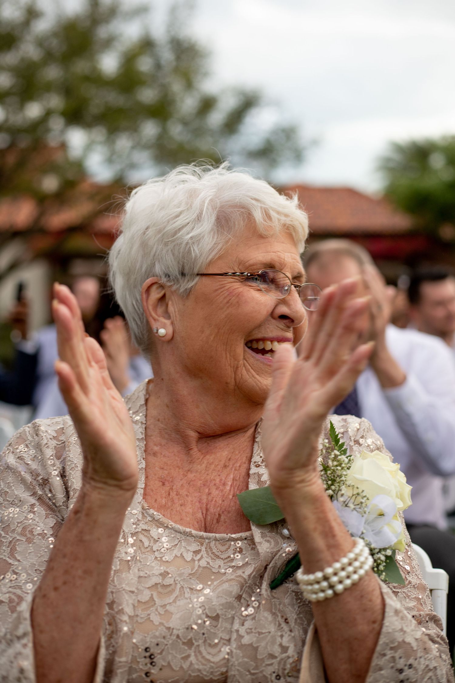Elderly woman in lace dress clapping and smiling joyfully at an outdoor wedding celebration.