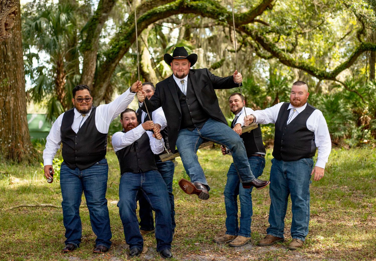 Groomsmen in western attire pose playfully under tree branches for wedding photos.