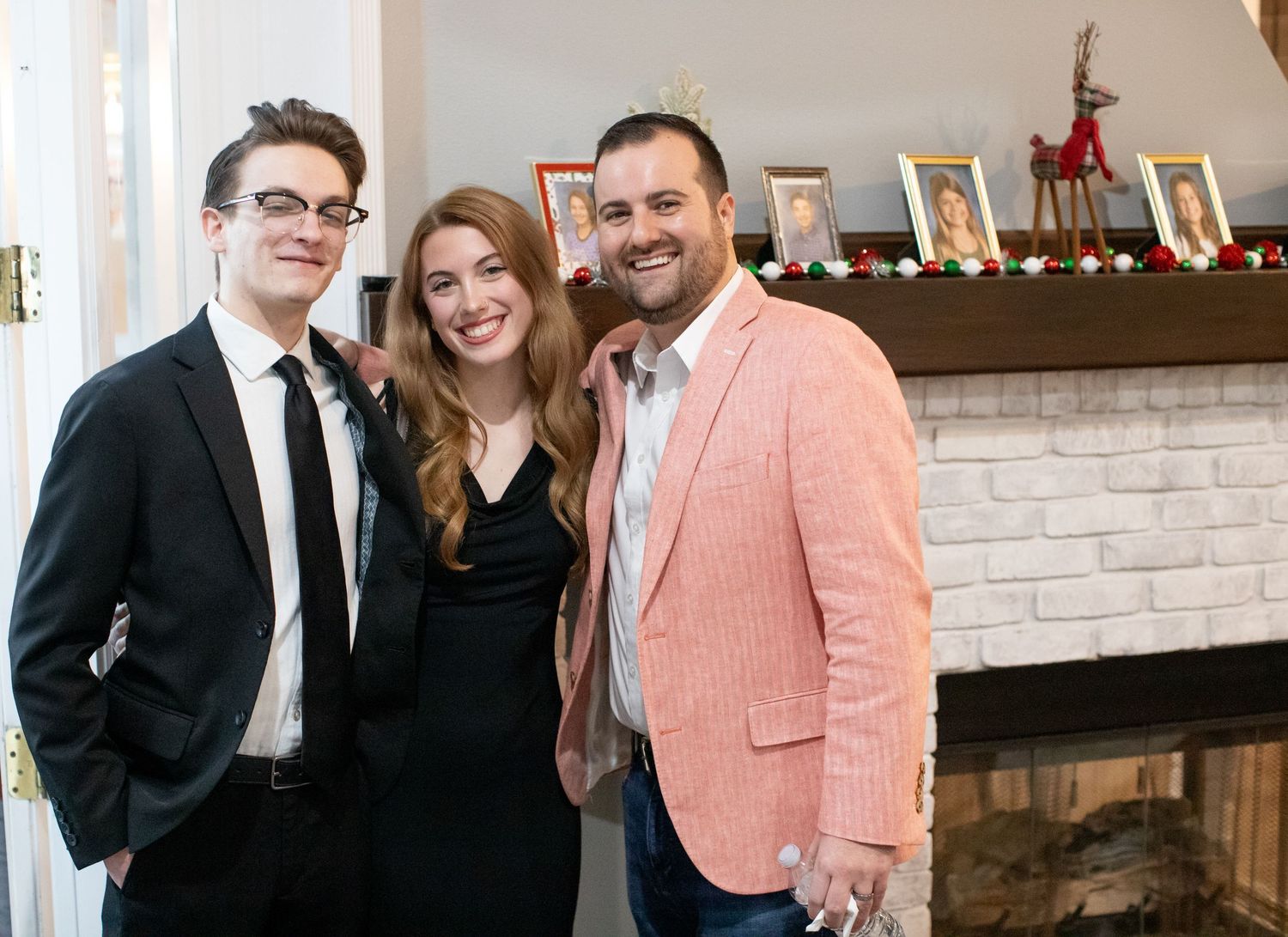 Group posing in formal attire by decorated fireplace at holiday gathering.