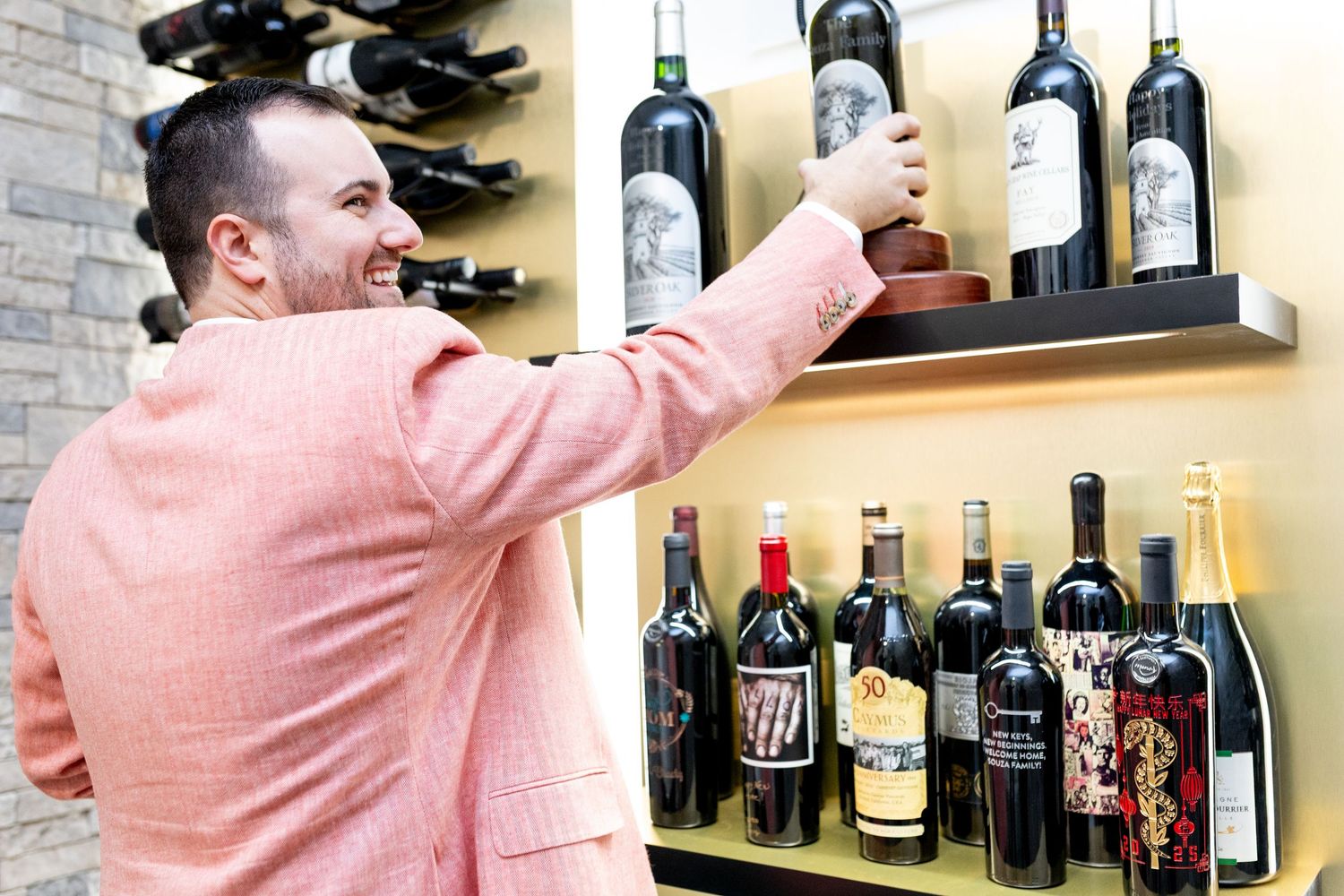 Person in salmon colored sweater reaching for wine bottles on display shelf.