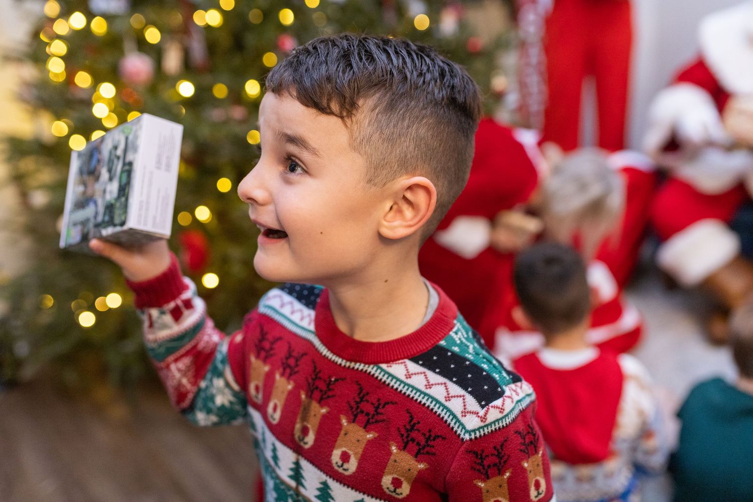 Children wearing festive sweaters celebrate during a holiday event with decorated trees.