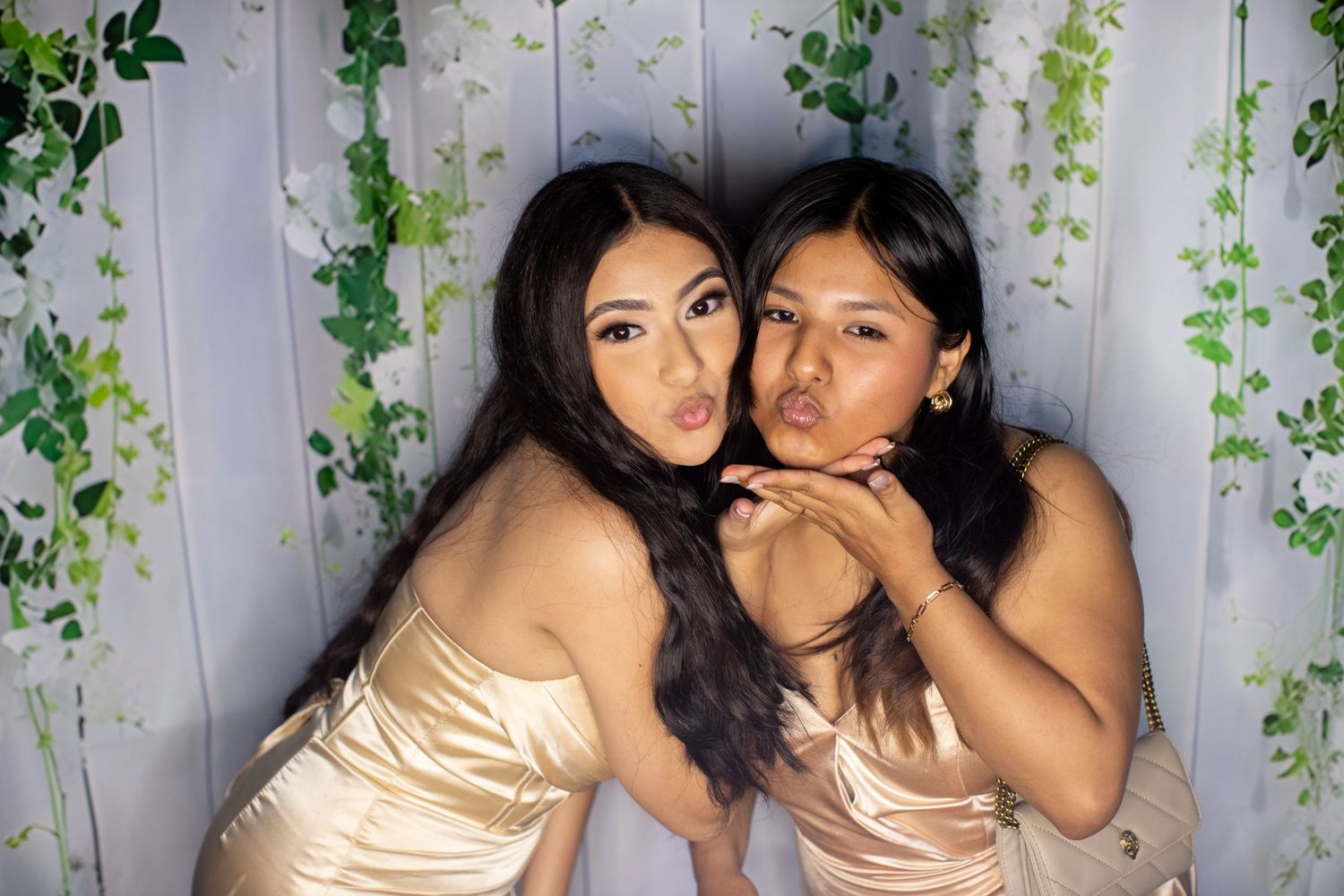 Two young women in champagne satin dresses posing together blowing kisses at a floral photo booth backdrop.