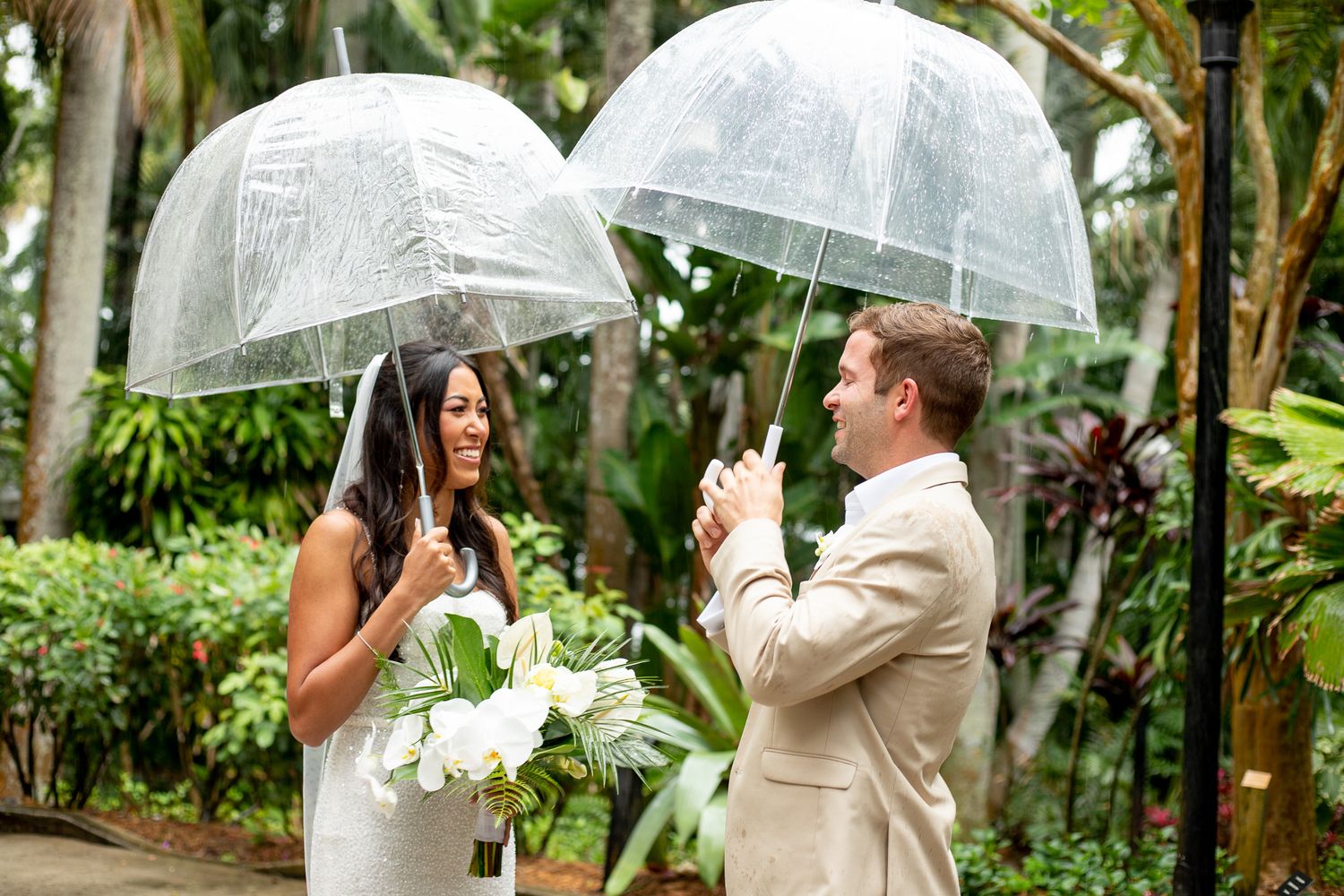 Bride and groom holding clear umbrellas during a rainy outdoor wedding ceremony surrounded by tropical greenery.