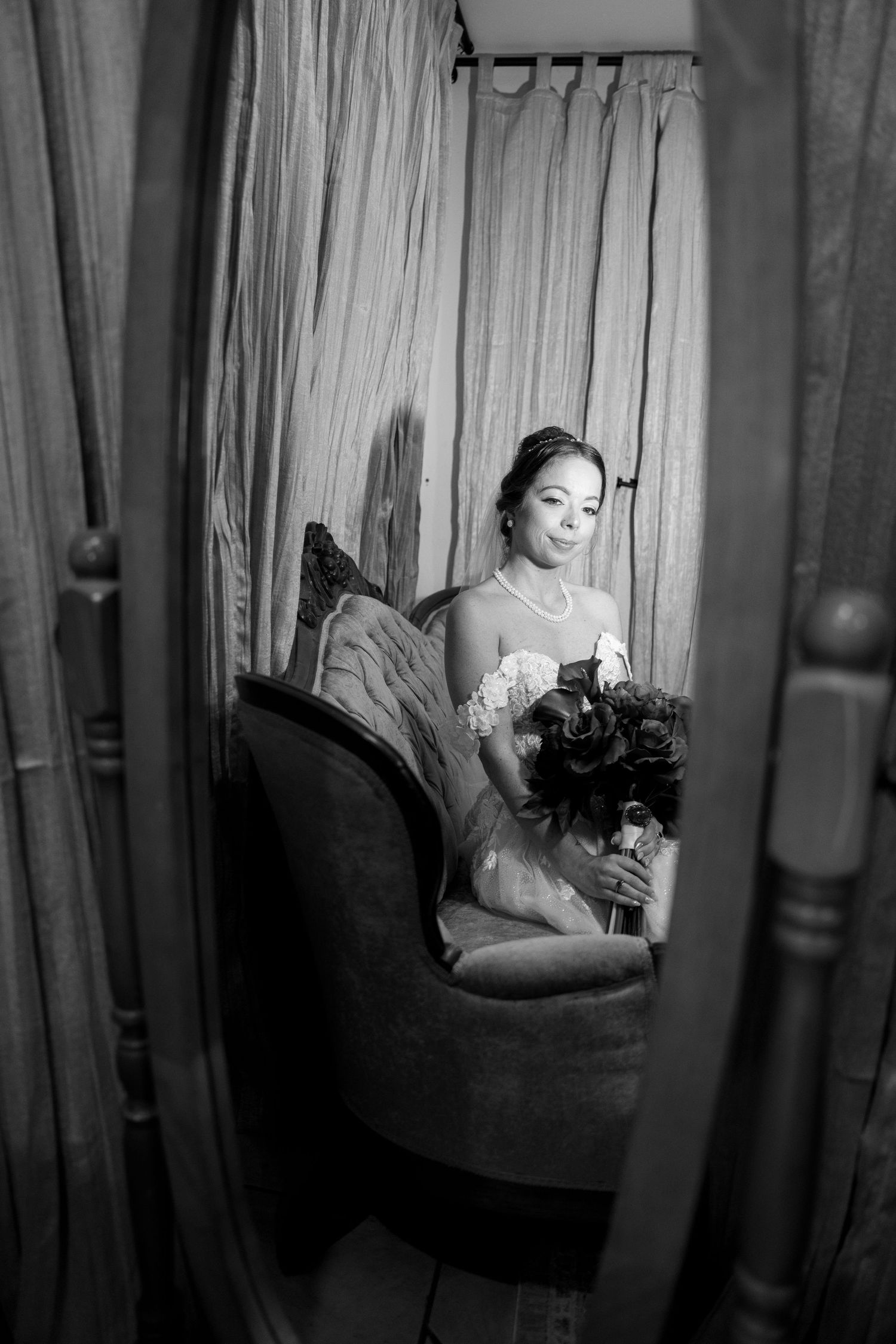 Black and white photo of a bride in a wedding dress holding a bouquet, seated on a vintage chair reflected in a mirror.