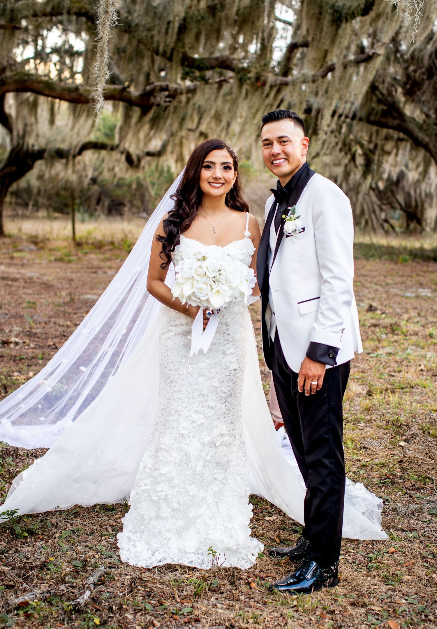 A bride in a lace gown with flowing veil poses in a natural outdoor setting.
