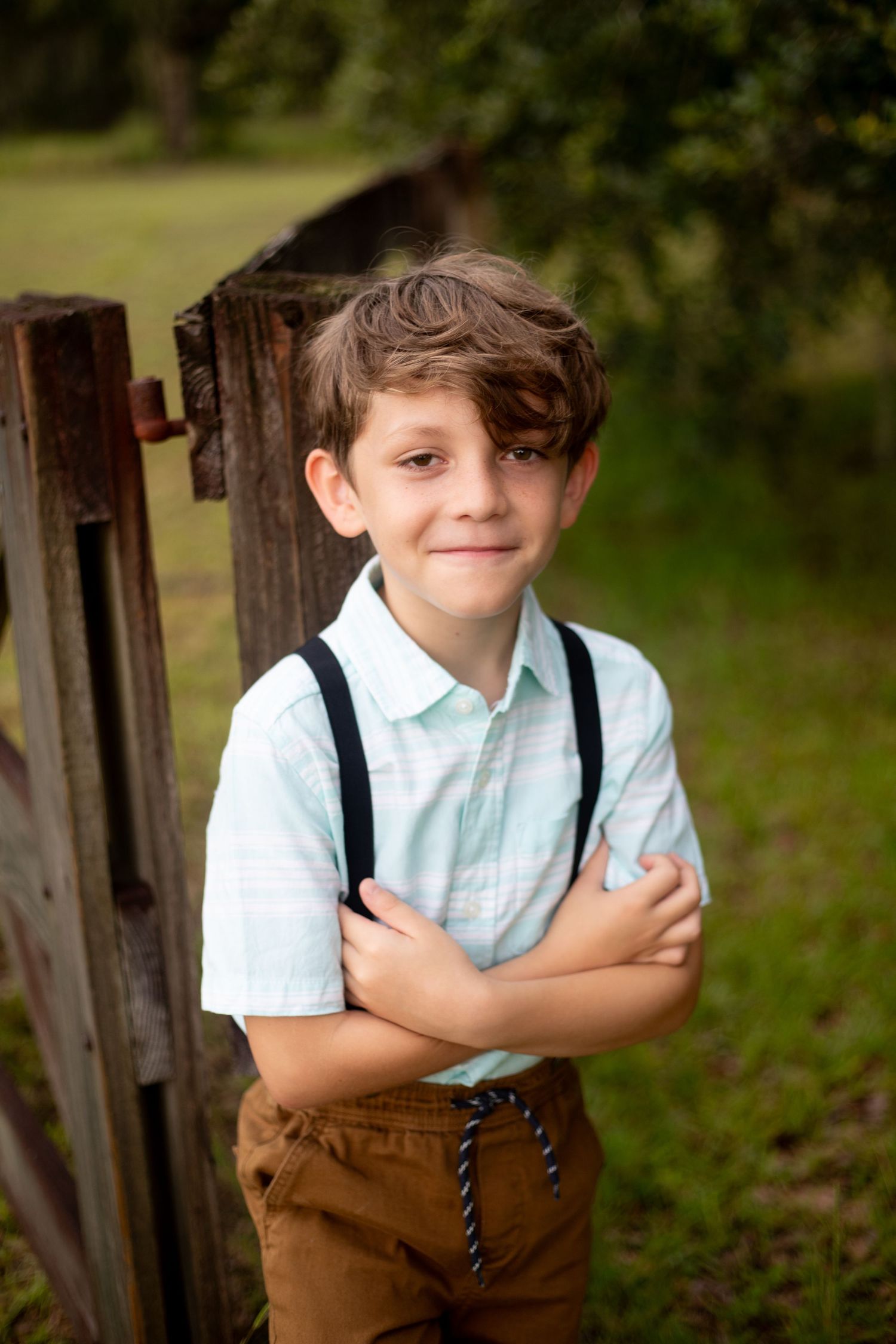 Child in vintage-style outfit with suspenders poses near wooden fence in outdoor setting.