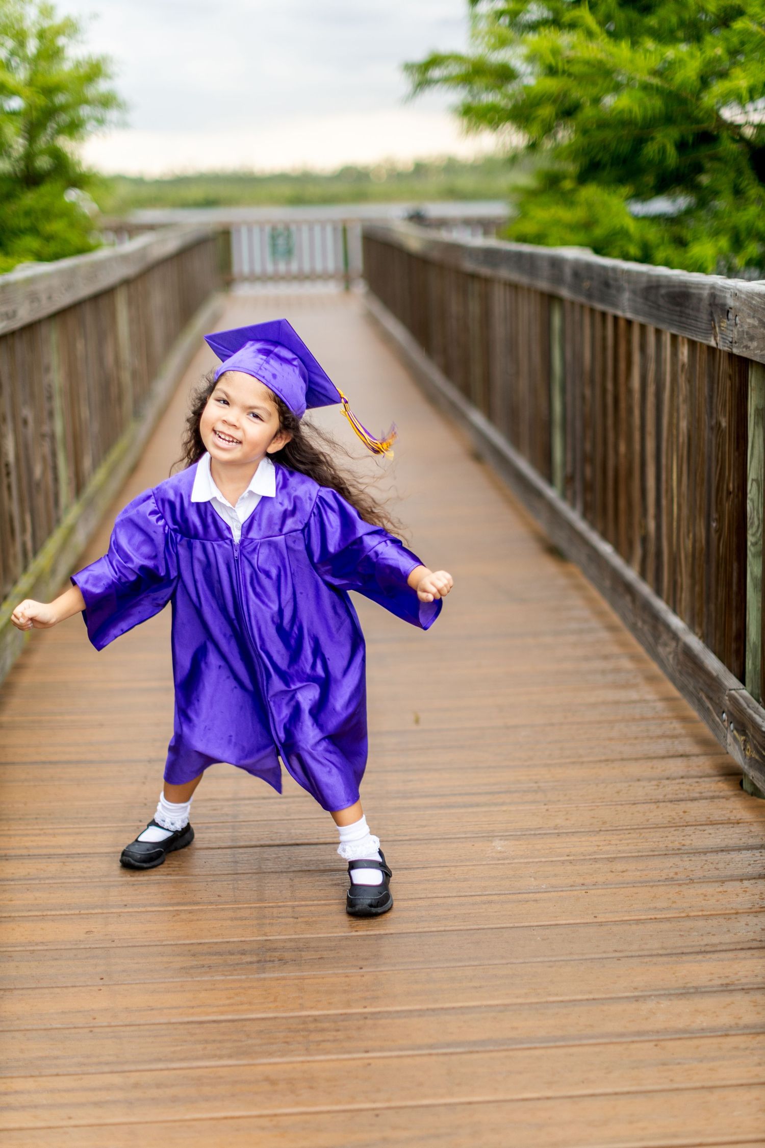 Young graduate in purple cap and gown playfully posing on a wooden boardwalk surrounded by greenery.