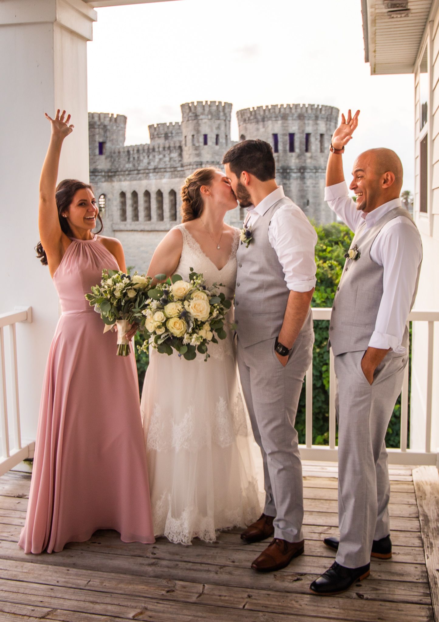 Wedding party in pink and grey attire poses on white balcony with castle towers visible.