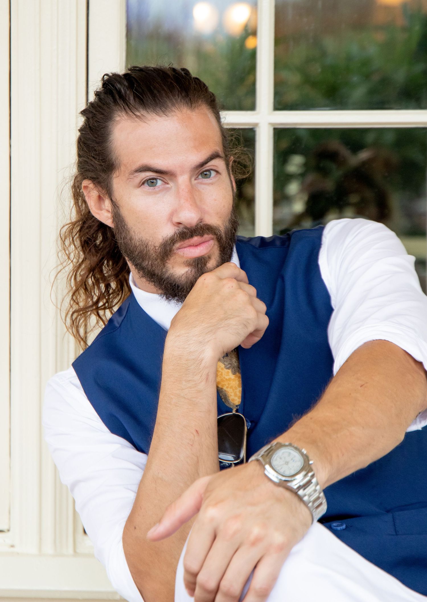 Person in blue vest and white shirt poses thoughtfully by window with sunglasses.