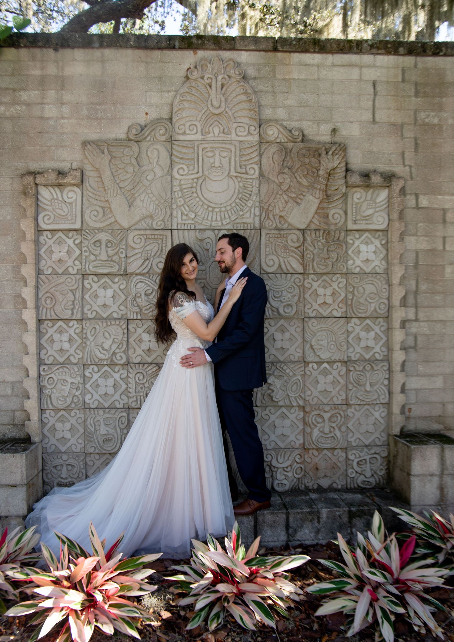 Bride and groom embracing before ornate stone wall with tropical plants at outdoor wedding venue.