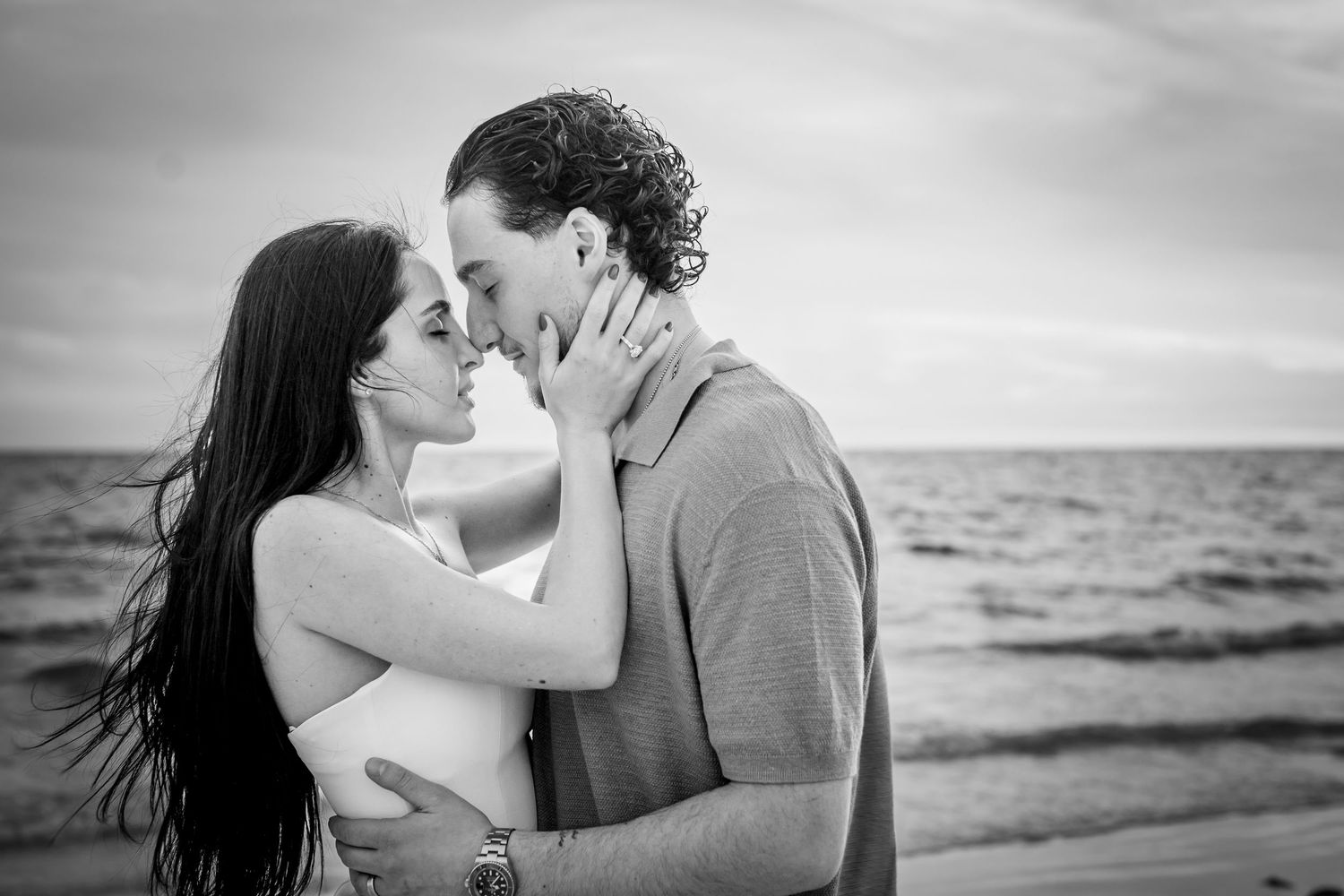 Black and white photo of a couple sharing a romantic kiss on a beach with ocean waves in the background.