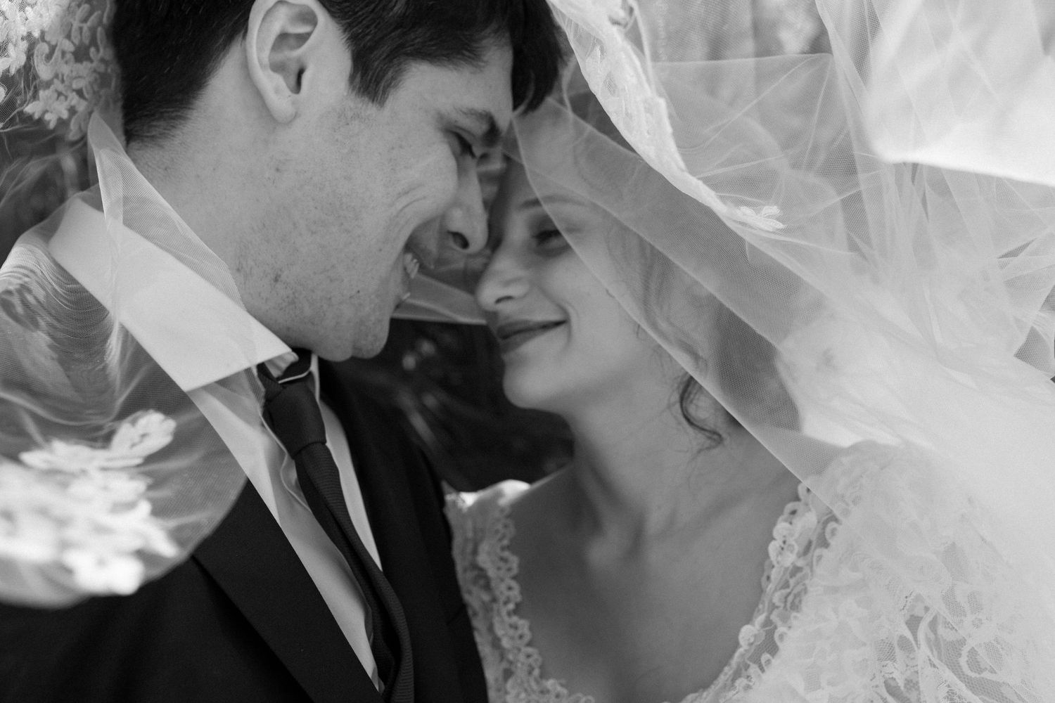 Romantic black and white photo of bride and groom smiling under wedding veil outdoors.