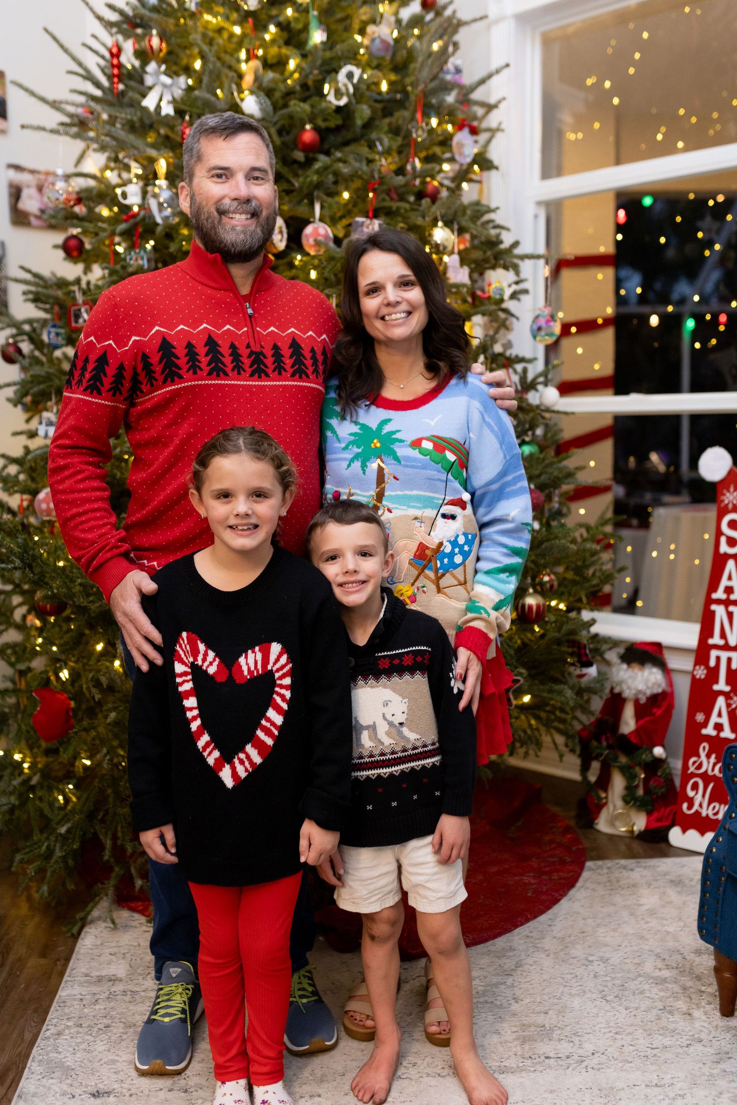 Family wearing festive holiday sweaters poses in front of decorated Christmas tree.