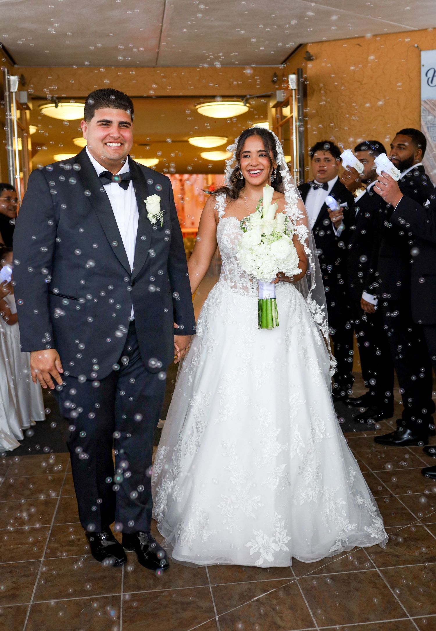 Bride and groom exit wedding venue surrounded by bubbles, bride in lace gown holding white bouquet, groom in black tuxedo.