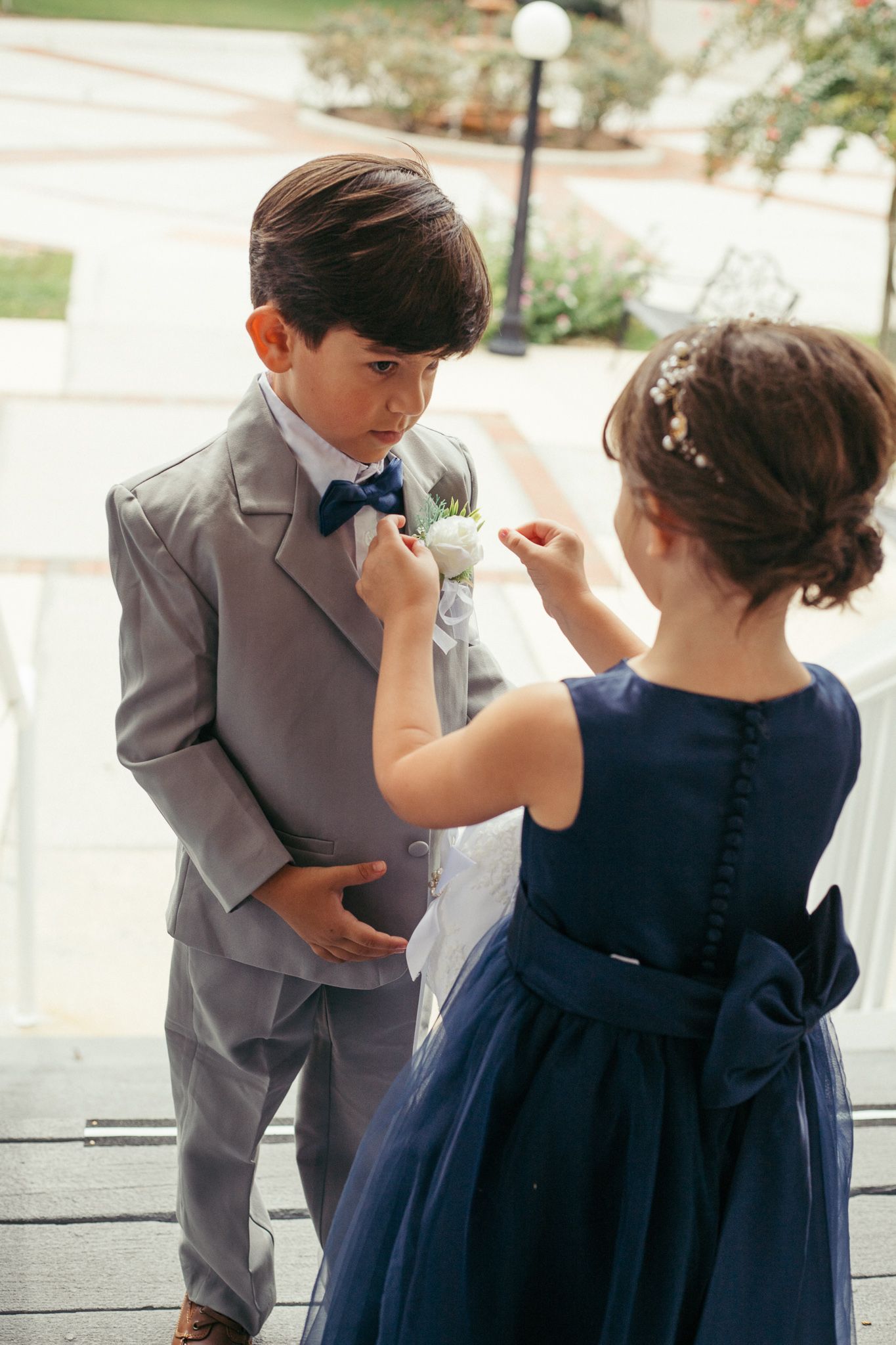 A young ring bearer in a gray suit and bow tie gets his boutonniere adjusted by a flower girl in a navy blue dress.