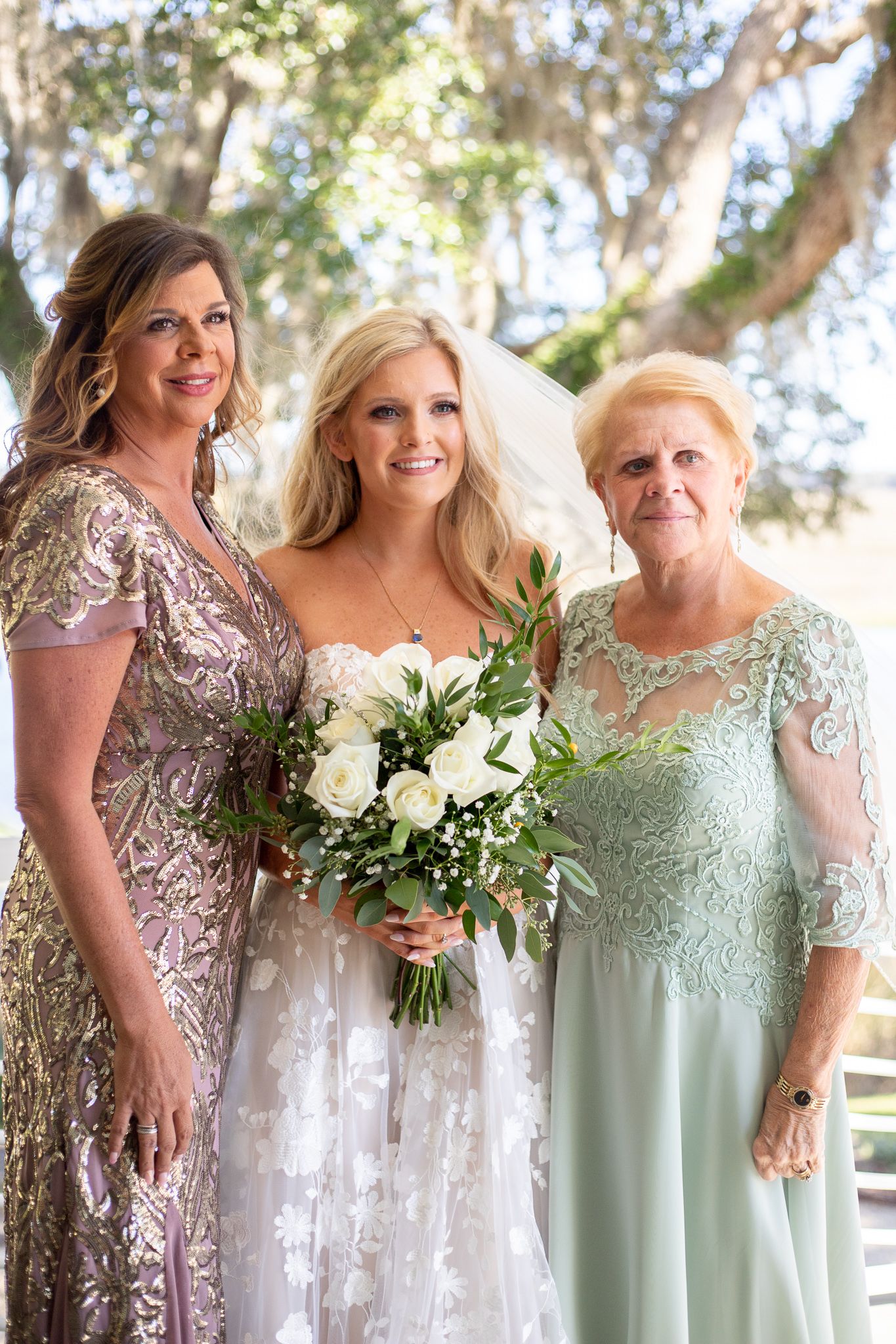 Bride in white gown holding bouquet poses with two women in elegant dresses at an outdoor wedding.