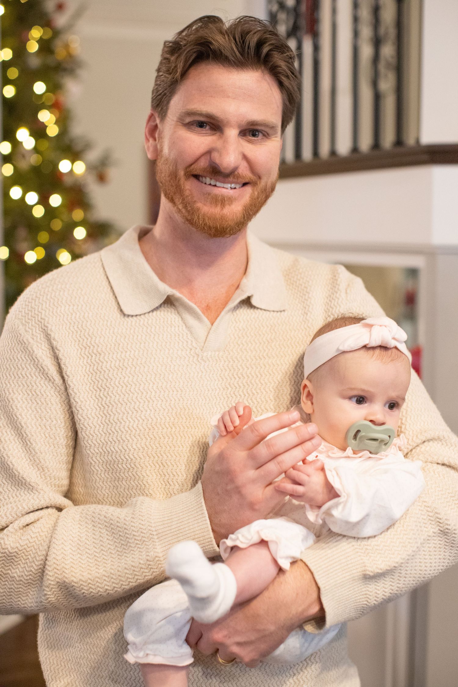 Person in cream sweater holding a baby near a Christmas tree with twinkling lights.