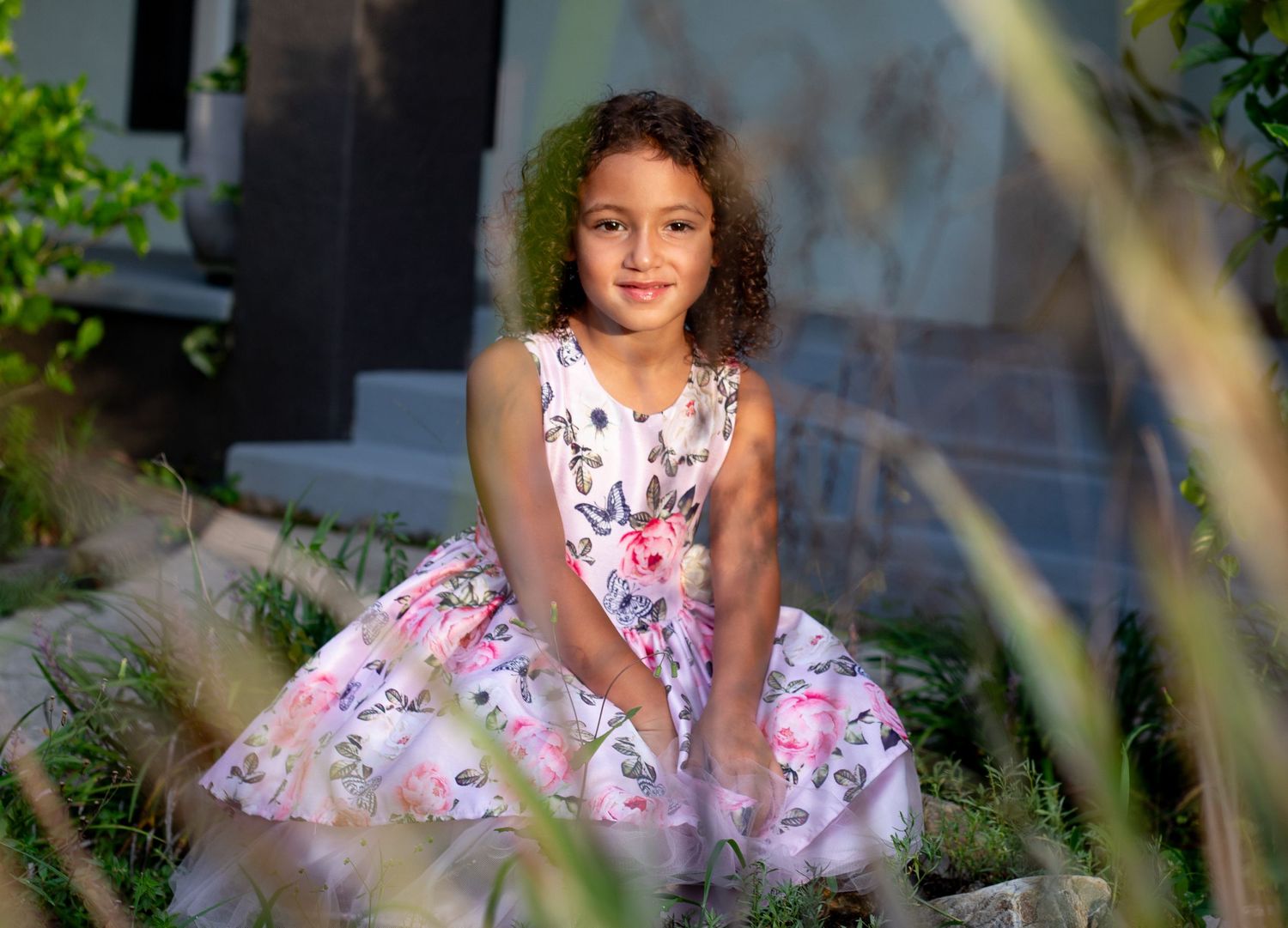 Child in pink floral dress poses among garden greenery and sunlit foliage.