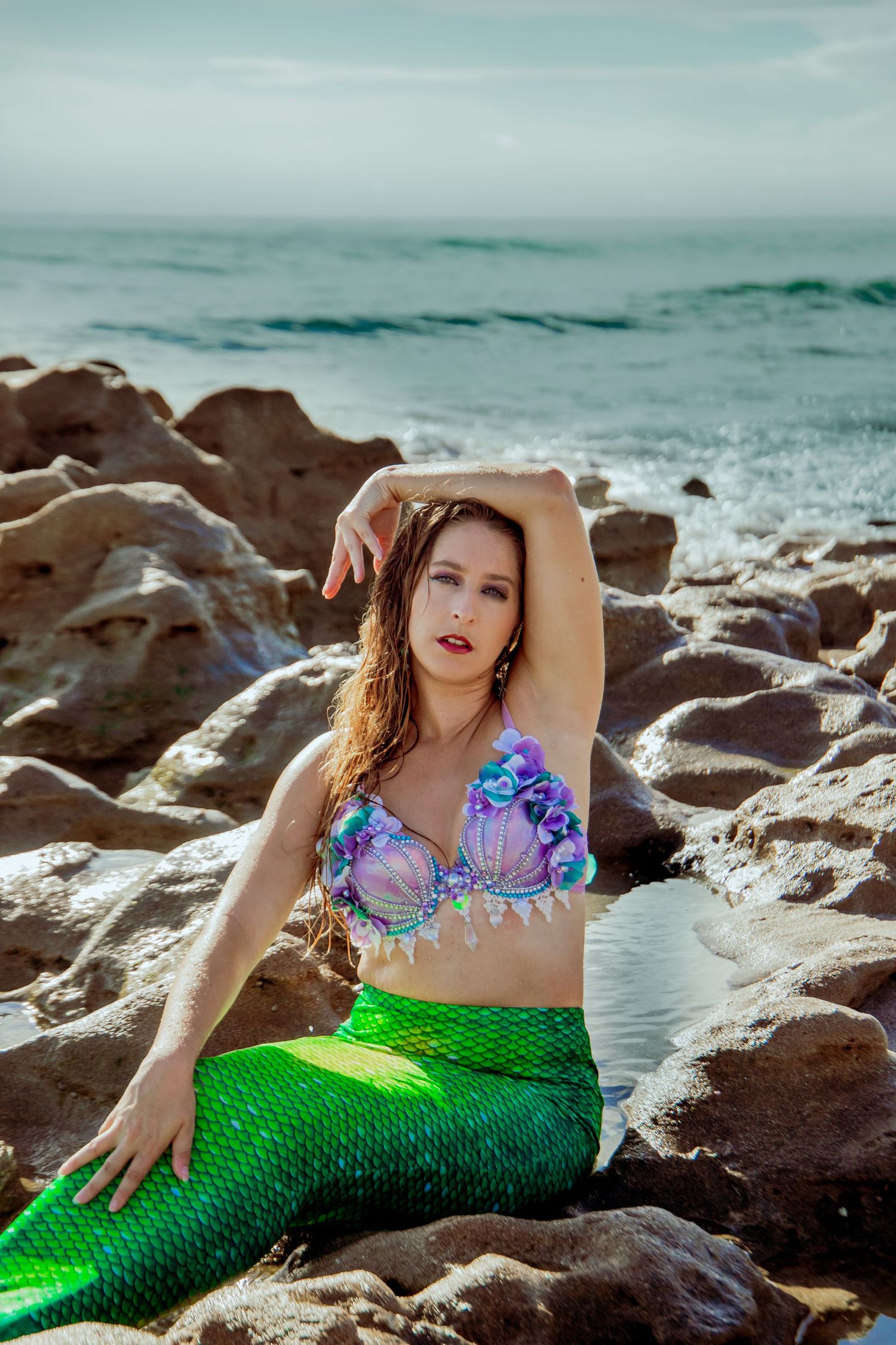 Mermaid-costumed model in purple top and green tail poses dramatically on coastal rocks.