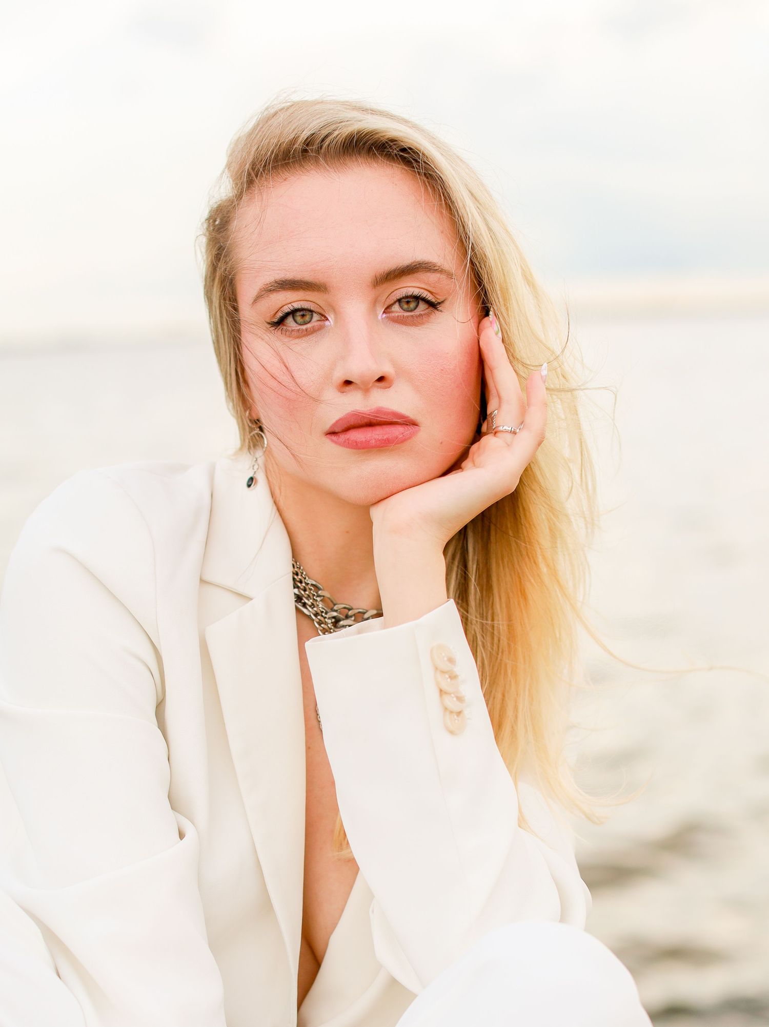 Soft portrait with statement necklace and white blazer against seaside backdrop.