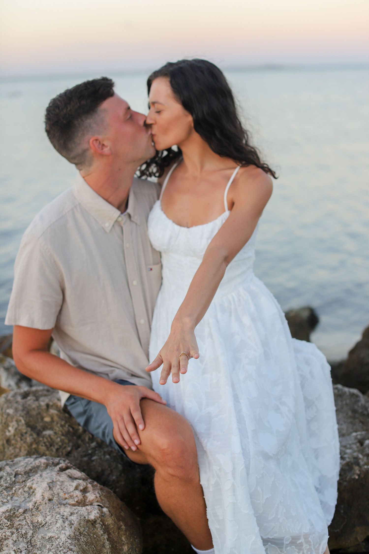 Couple kissing on rocks by the ocean at sunset, woman in white dress and man in beige shirt.