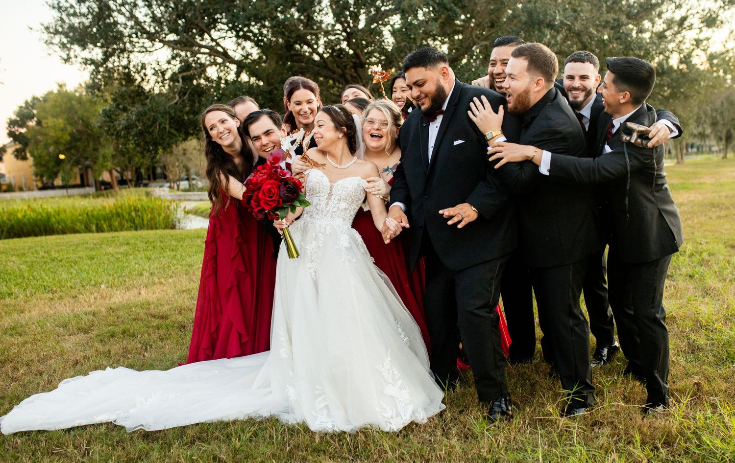 Wedding party in burgundy and black attire laughing together outdoors, bride in white gown with groom at center.