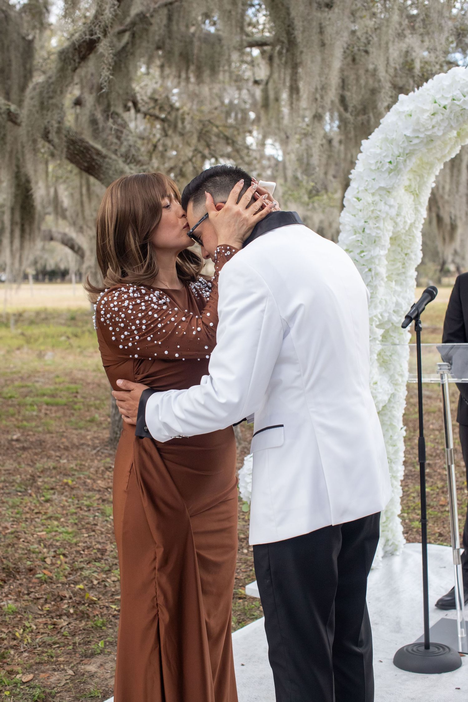 Couple sharing first kiss at outdoor wedding ceremony near white floral arch and moss-draped oak trees.