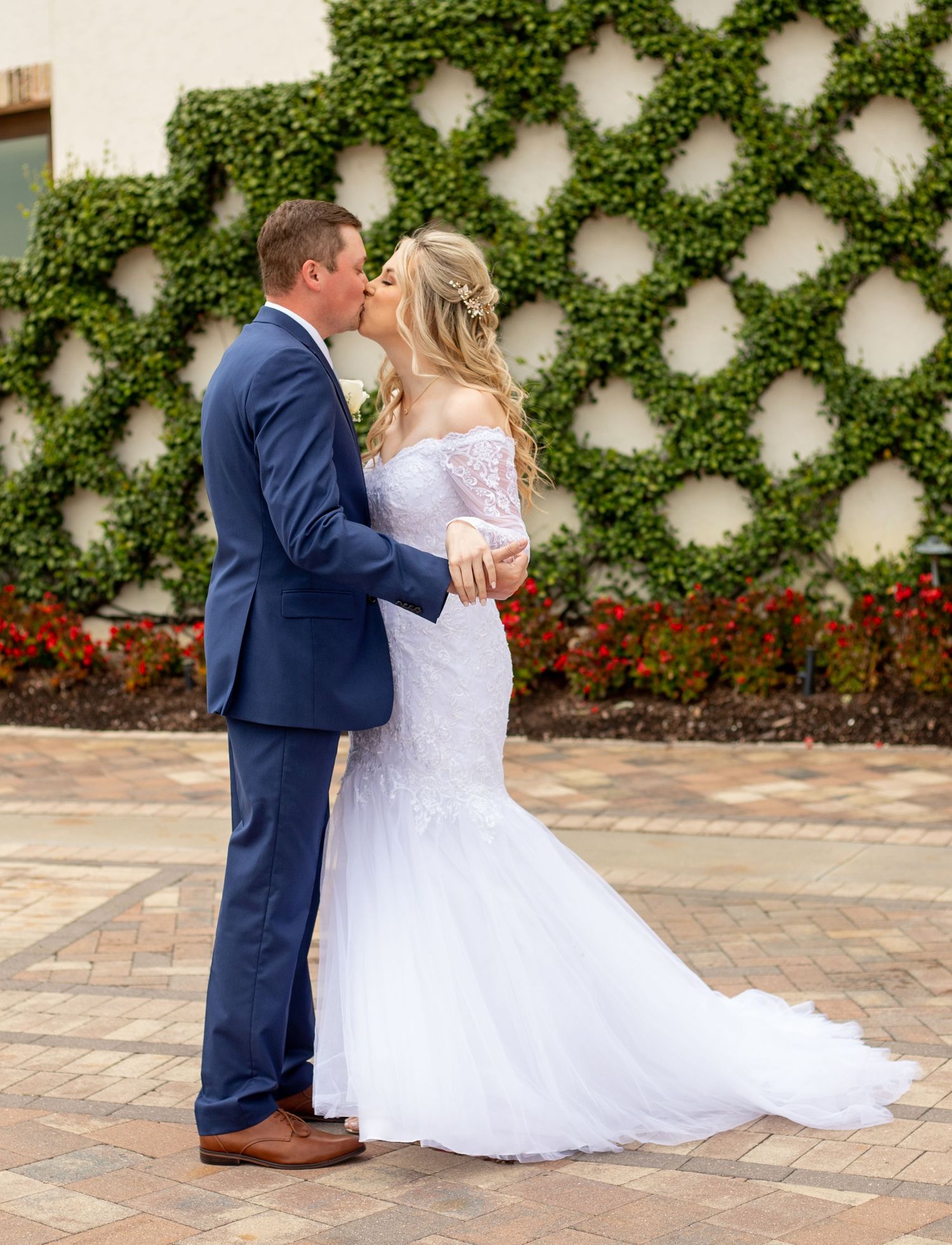 Bride and groom share a romantic kiss in front of an ivy-covered wall, bride in lace mermaid gown, groom in navy suit.