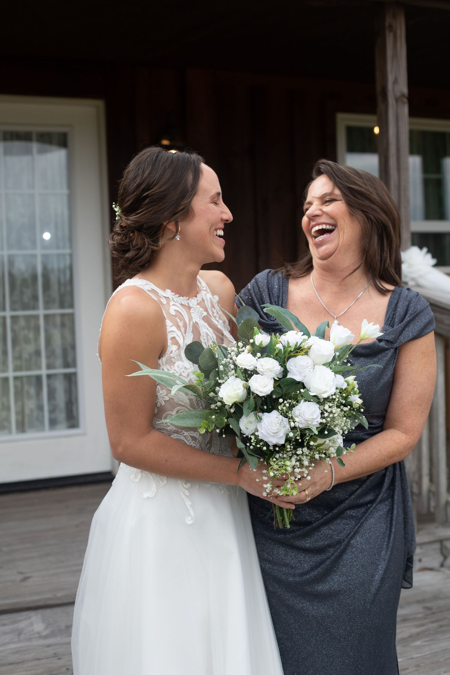 Bride in white lace gown laughing with woman in gray dress holding white rose bouquet on wooden deck.