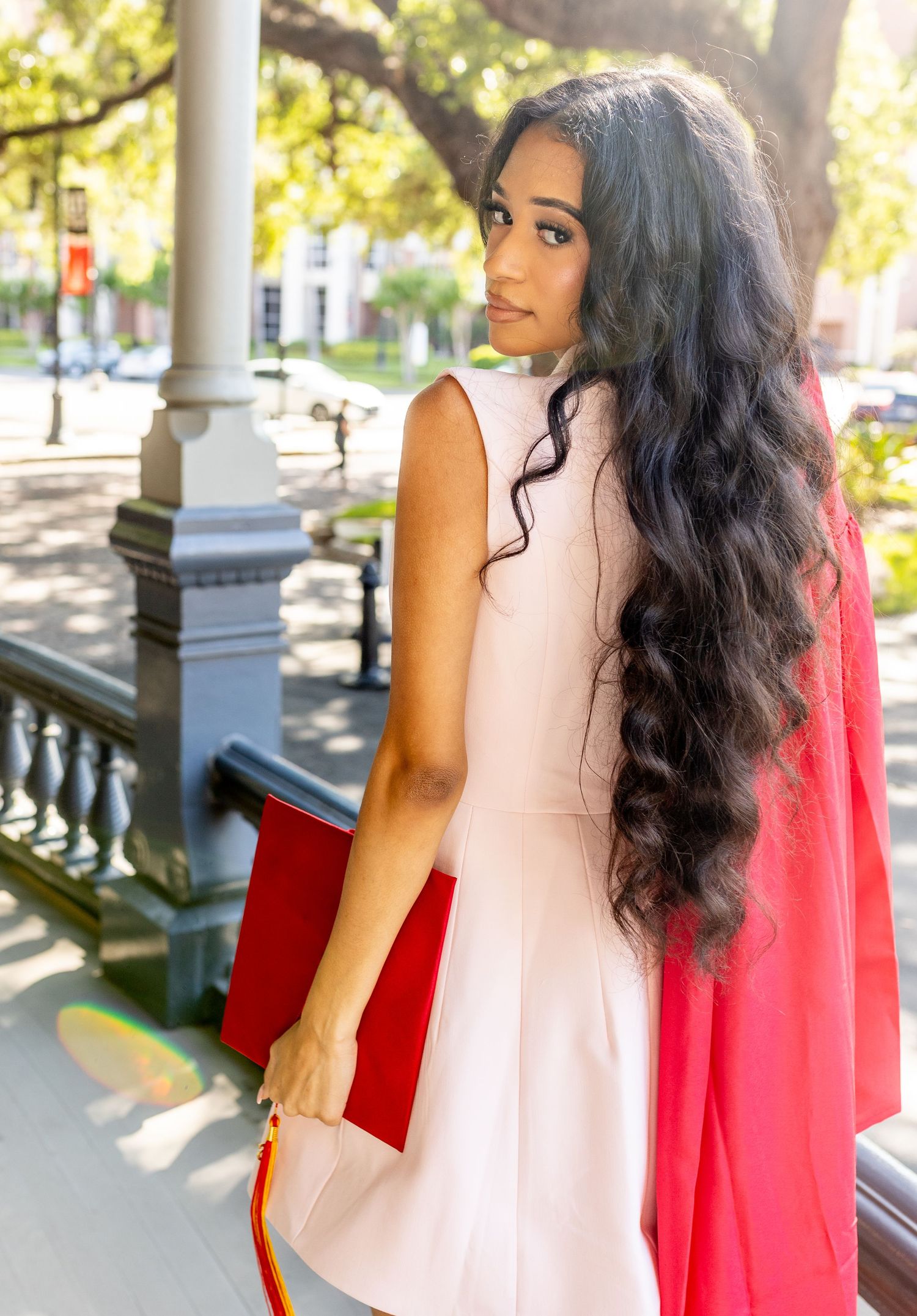 Young woman in pink dress with long curly hair holds red clutch, posing outdoors near white columns.