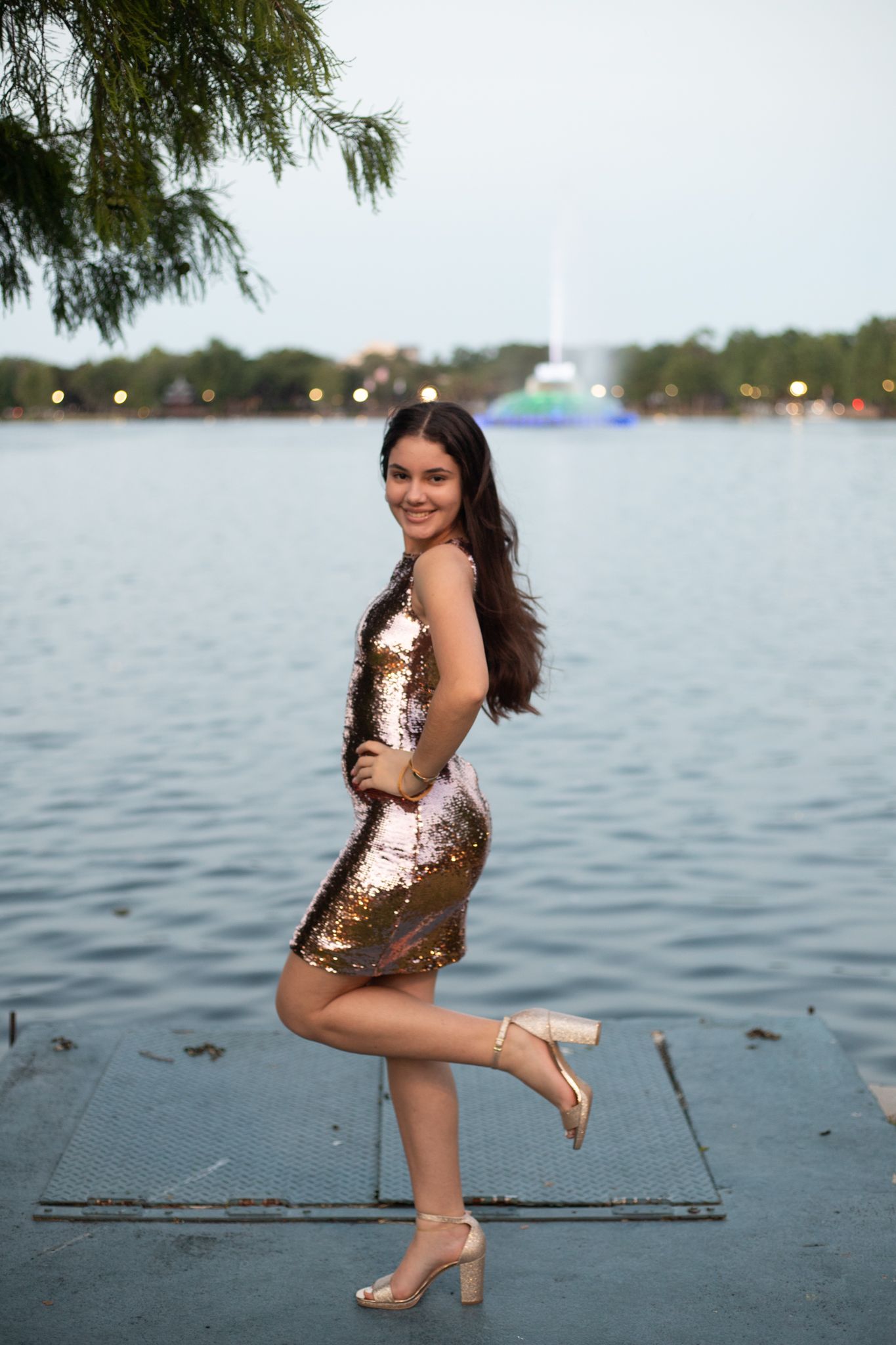 Celebratory pose in sparkly gold dress by waterfront with city skyline in background.