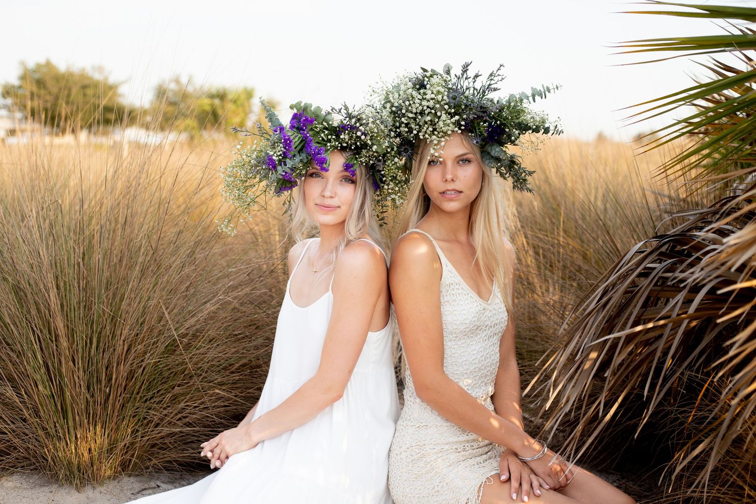 Two women in white dresses with flower crowns posed back to back outdoors.