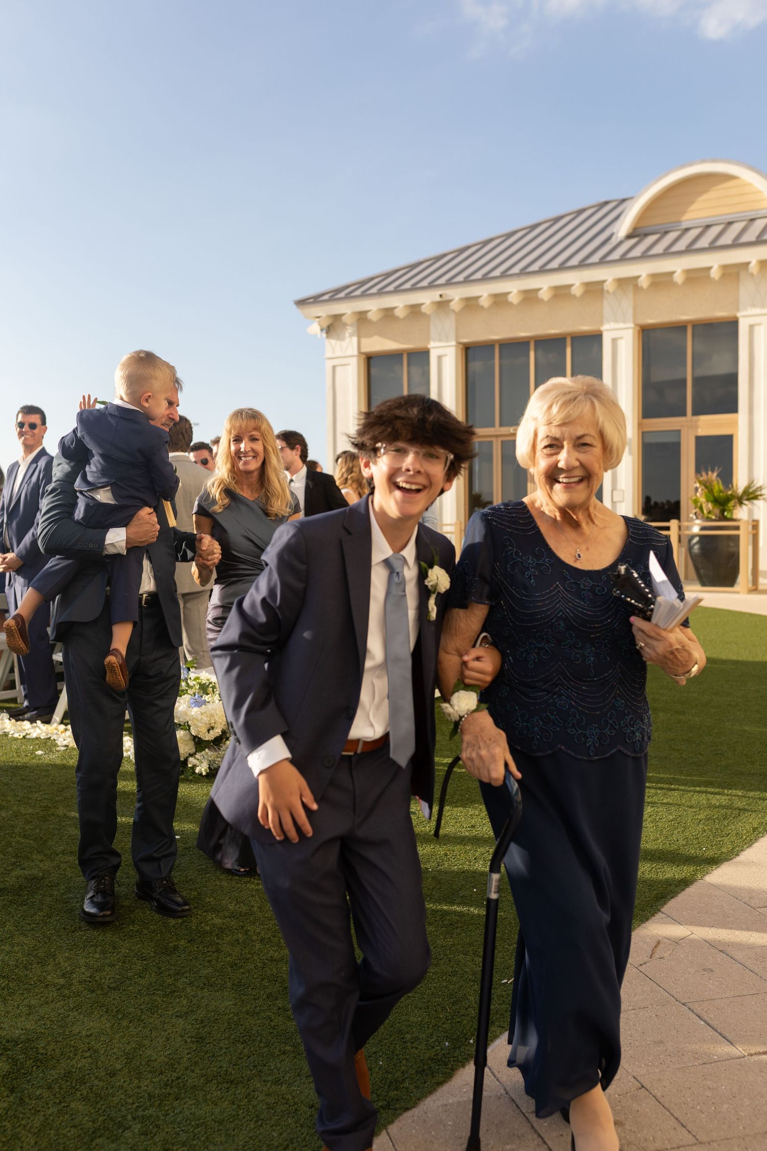 A young boy in a navy suit escorts an elderly woman in a blue dress at an outdoor wedding ceremony.