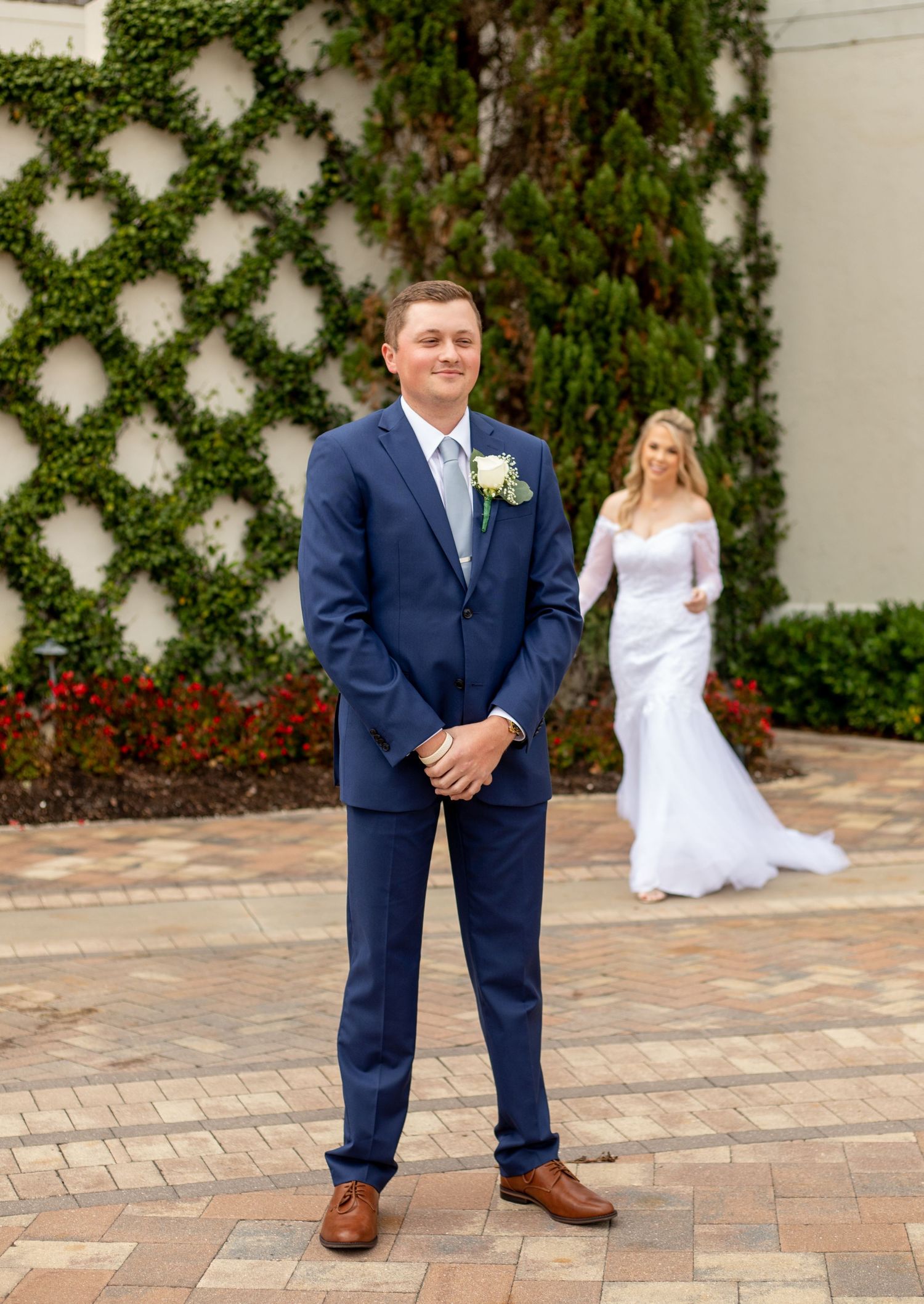 Groom in navy suit with boutonniere stands on brick patio as bride in white gown approaches from behind.