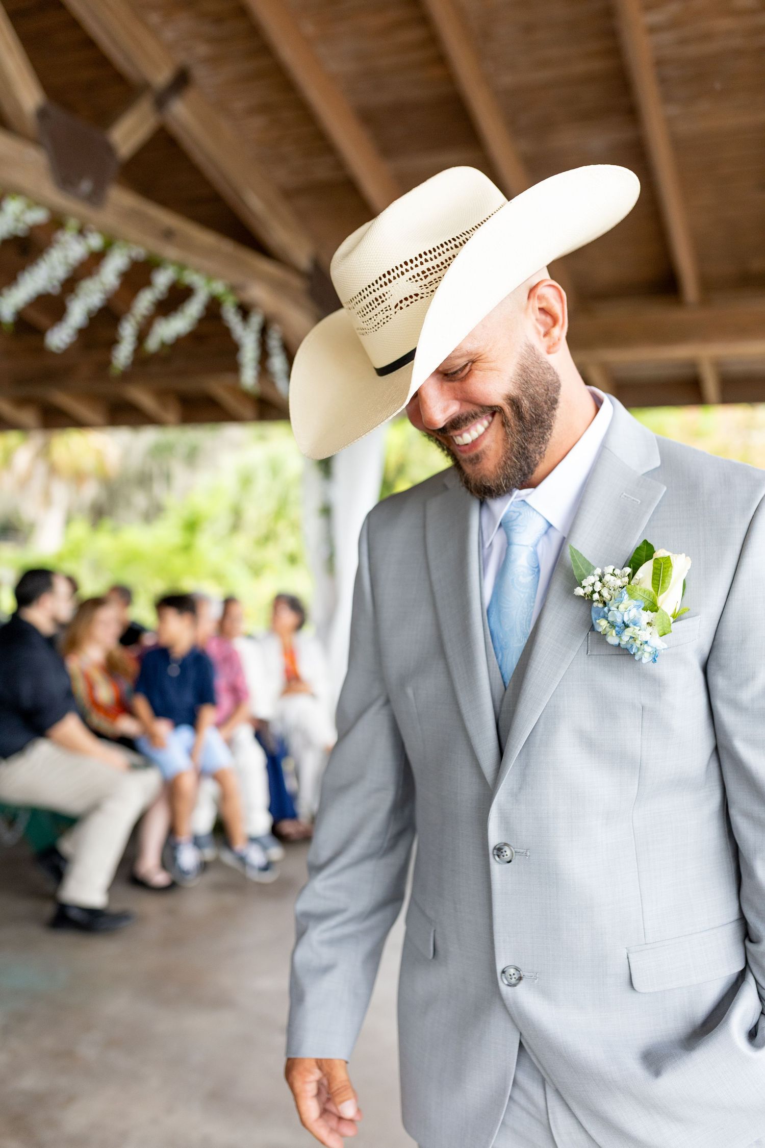 Smiling groom in gray suit, cowboy hat, and boutonniere walks at outdoor wedding ceremony with seated guests in background.