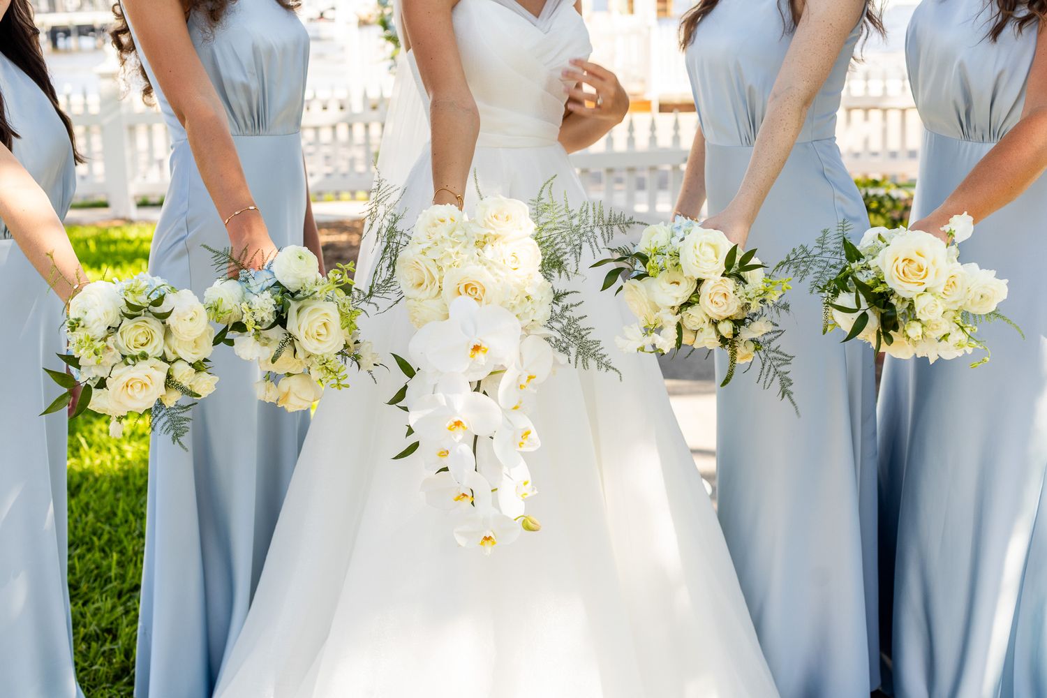 Bride in white gown with bridesmaids in light blue dresses holding white floral bouquets with orchids and roses outdoors.