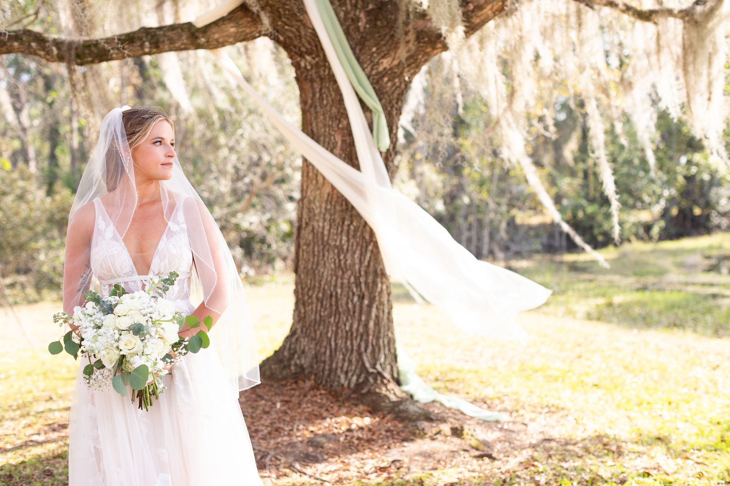 Bride in white gown poses under Spanish moss covered trees in natural outdoor setting.
