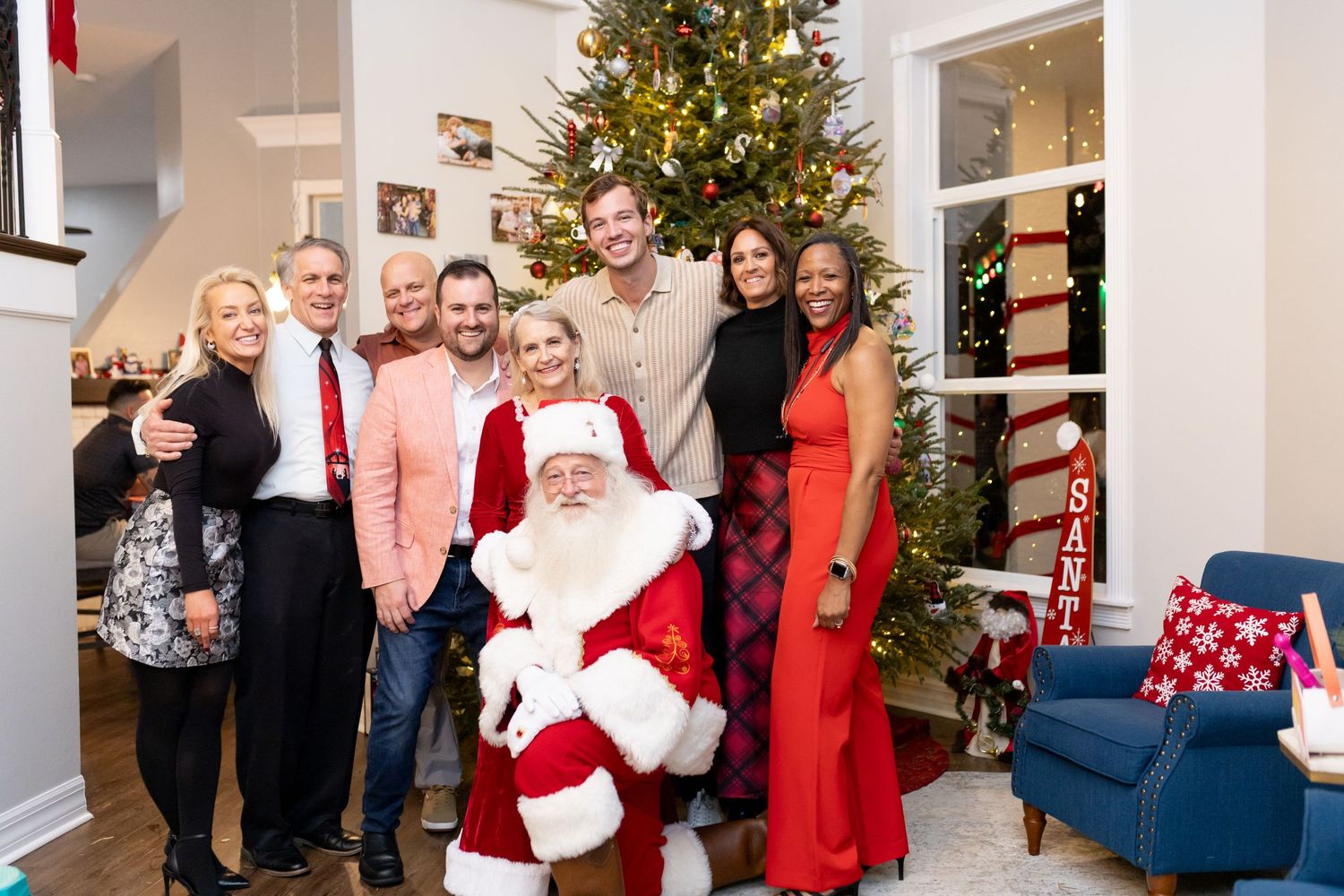 A festive holiday gathering with a decorated Christmas tree and people in formal attire in a bright interior space.