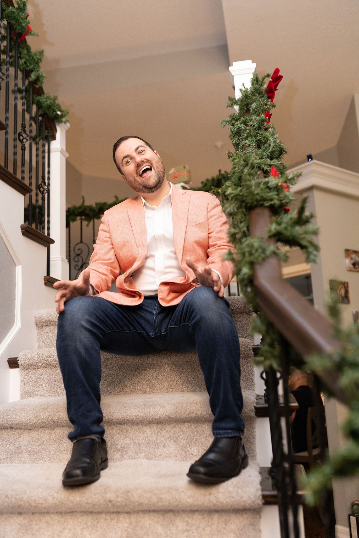 Someone wearing a peach-colored blazer and jeans sits on carpeted stairs decorated with holiday greenery.