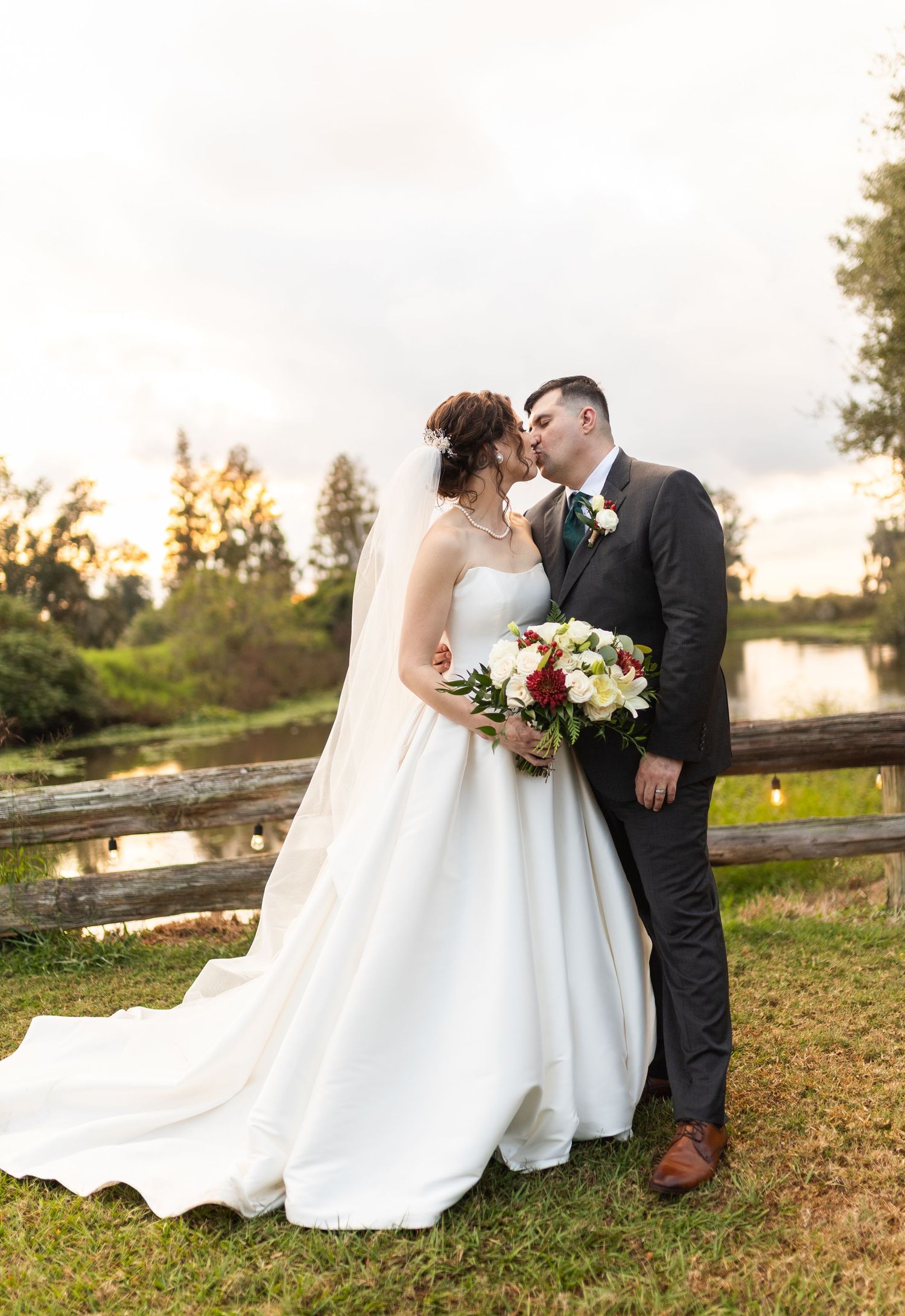 Romantic sunset wedding photos showing couple sitting on grass near water with flowing white dress.