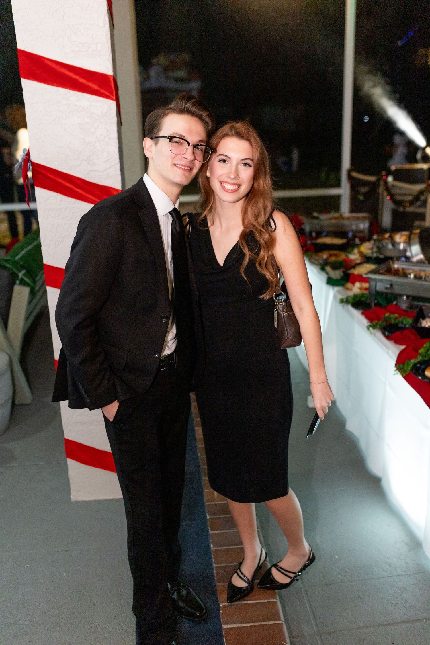 A couple in formal attire poses at an evening holiday party with festive decorations.