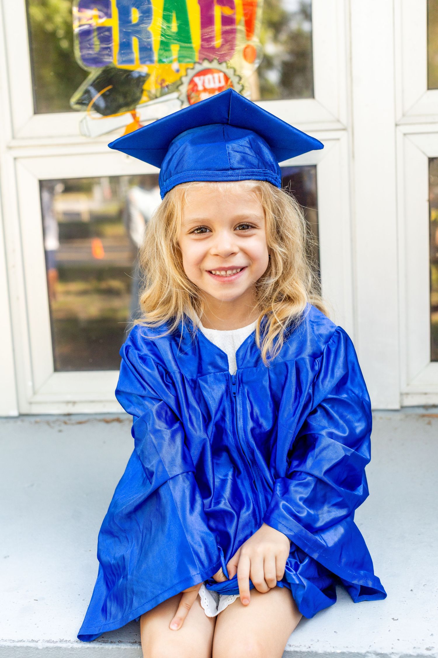 Graduate in blue cap and gown sits on steps with colorful graduation decorations in background.