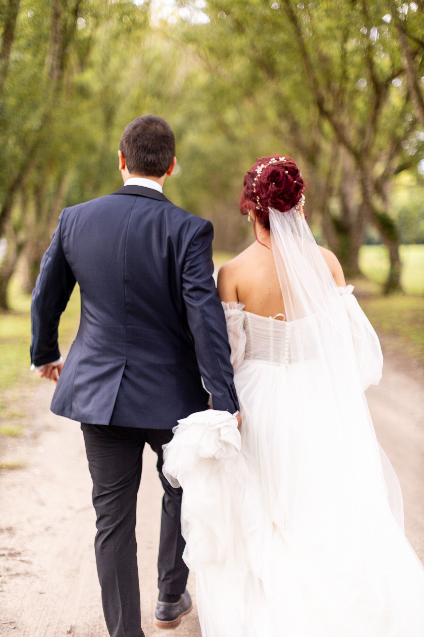 Bride and groom walking hand in hand down a tree-lined path, bride wearing a white gown with red floral hair accessories.