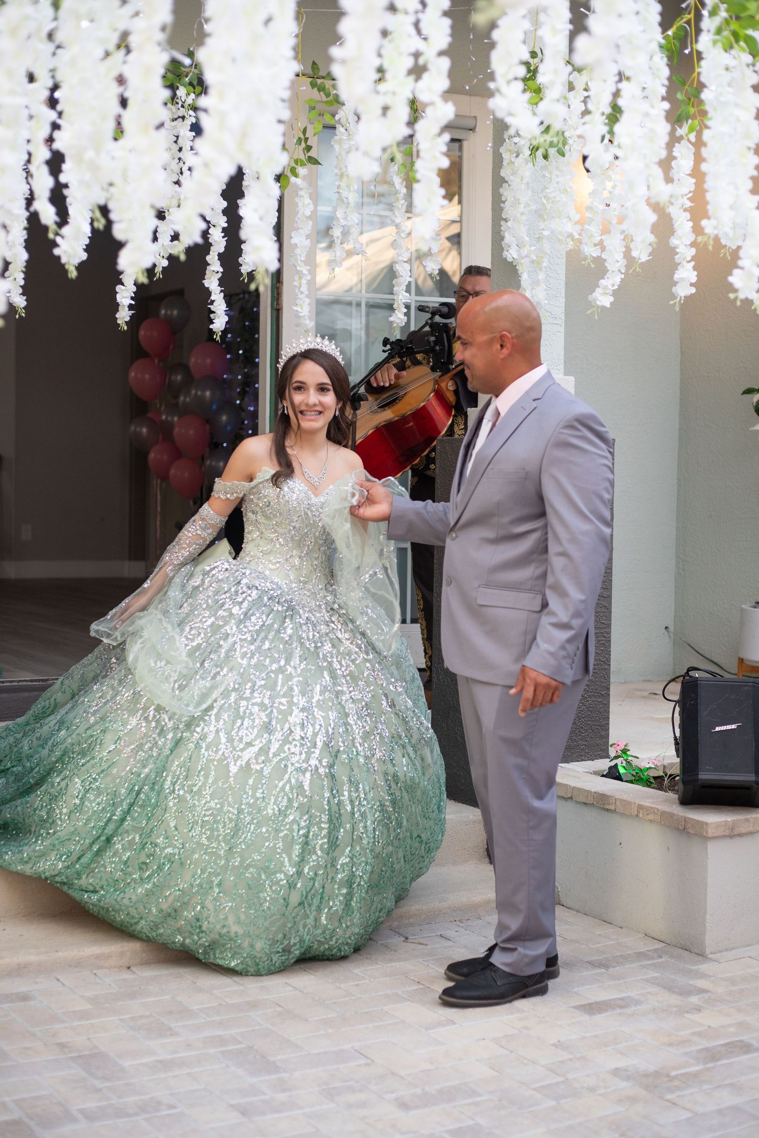 Person in a glittering mint ball gown posing under hanging white floral decorations.