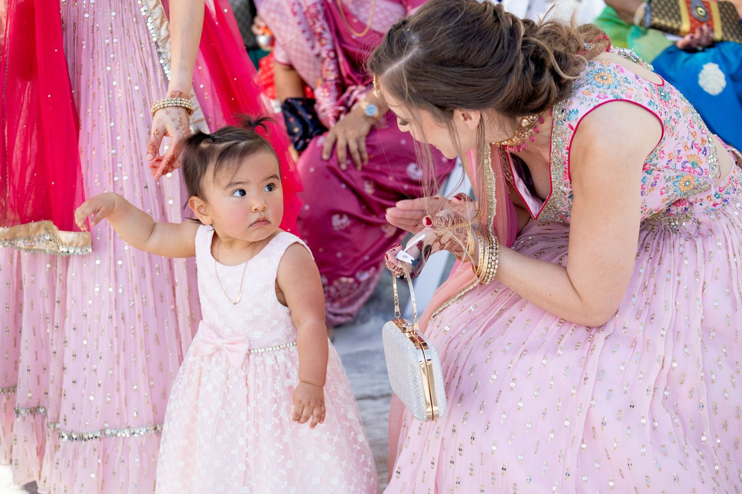 A toddler in a white dress interacts with a woman in a pink lehenga at a vibrant Indian celebration.