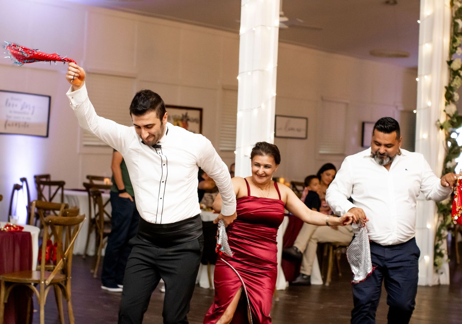 Three guests dance joyfully at a wedding reception, with a man in white raising a red accessory.