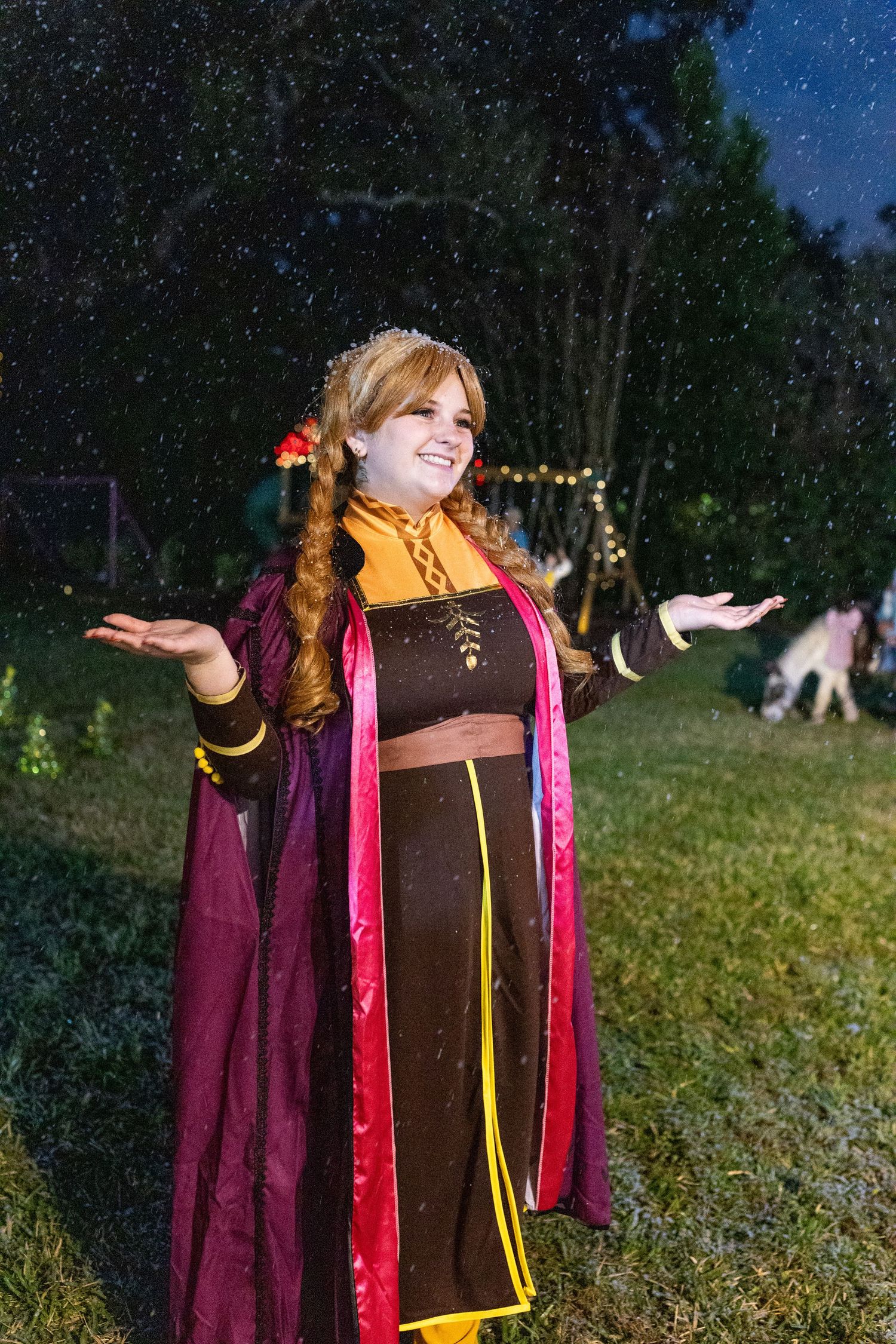 Person in a burgundy cloak poses playfully outdoors at night with twinkling lights in the background.