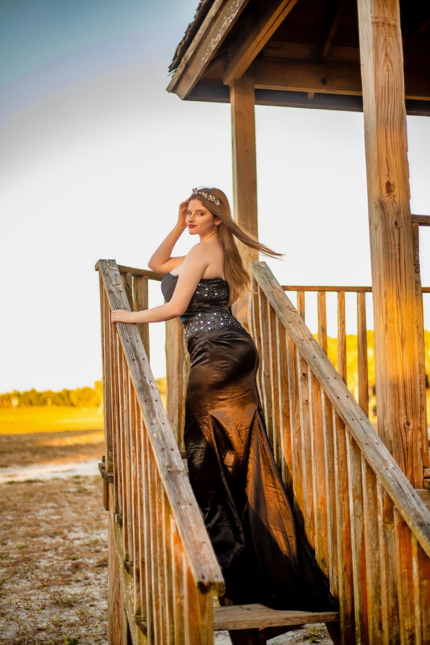 Dramatic portrait on wooden deck stairs in black evening gown at sunset.