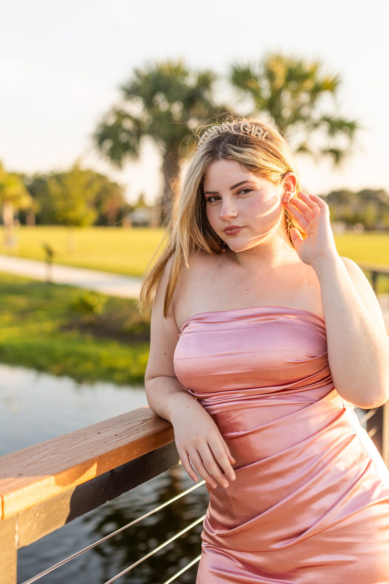 Pink satin dress glowing in golden sunset light by a waterfront railing.